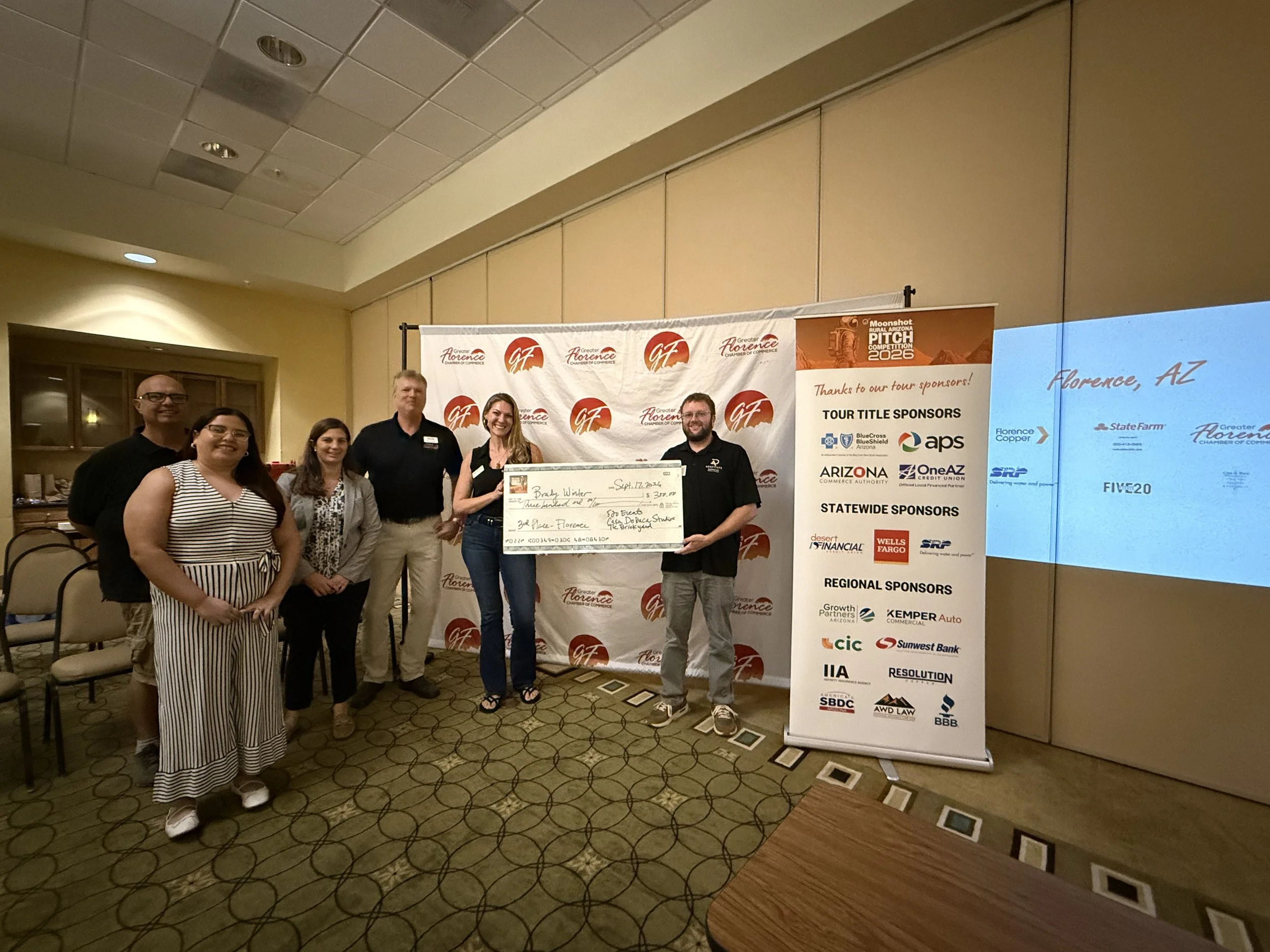 Six people standing together on a patterned carpeted floor in front of a backdrop with logos, holding a large check, at an indoor event in Florence, Arizona. The backdrop contains logos of sponsors and the event title, indicating a business or community gathering.