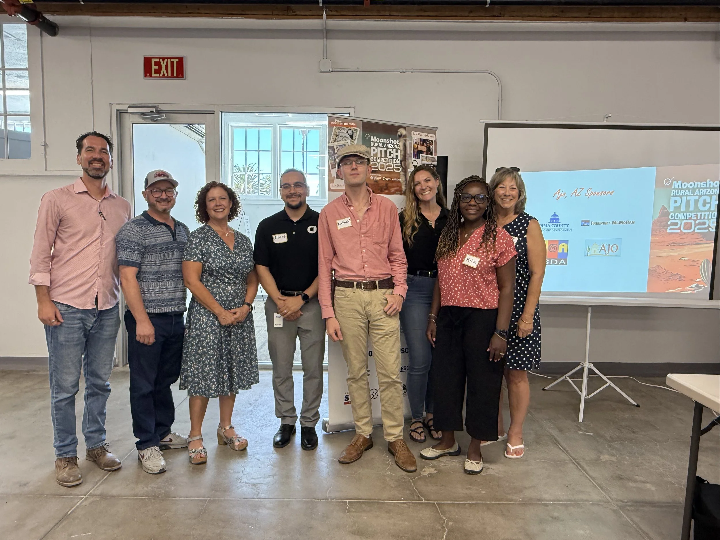 Group of nine diverse people standing indoors at a presentation event, with a projection screen behind them displaying logos and text for the Moonshot Rural Arizona Pitch Competition 2023.