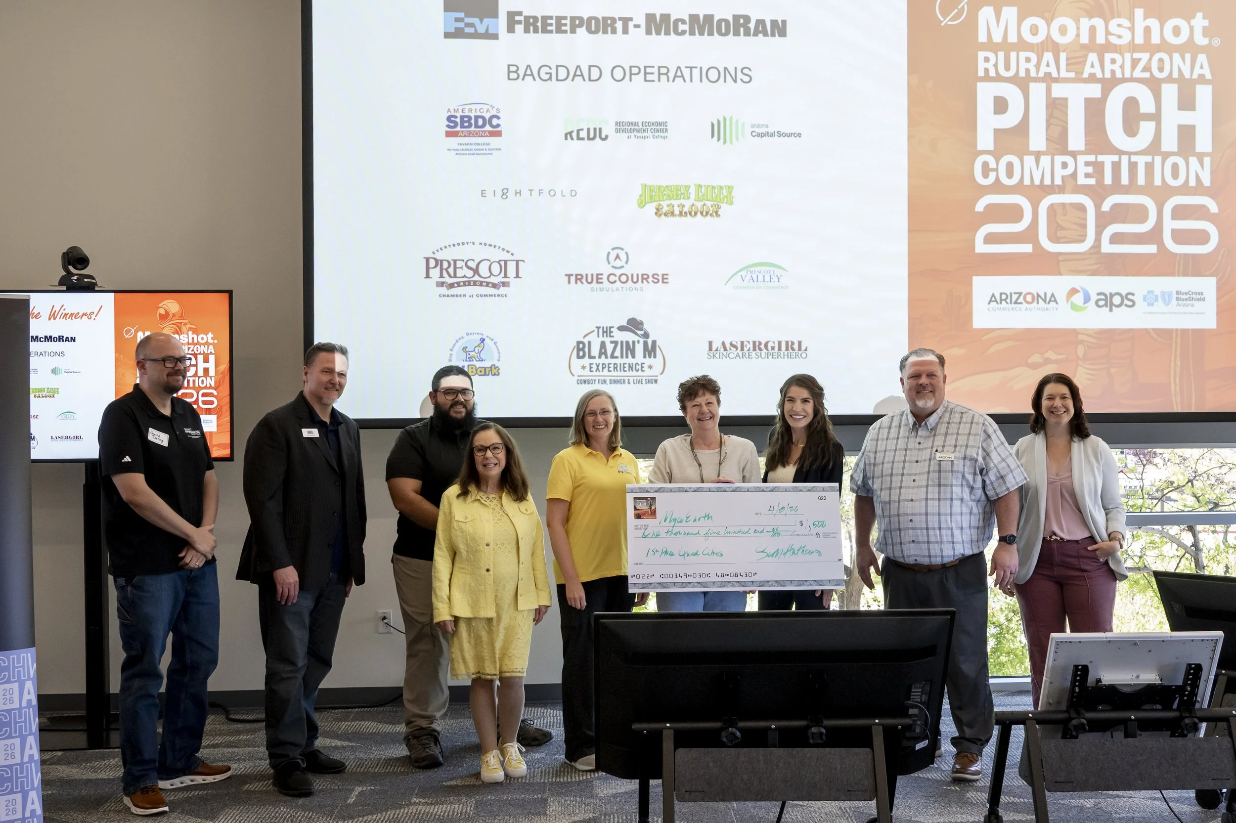 Group of people standing with oversized check in front of large screen displaying logos and event information for the 2026 Moonshot Rural Arizona Pitch Competition, with individuals smiling and posing for the photo.
