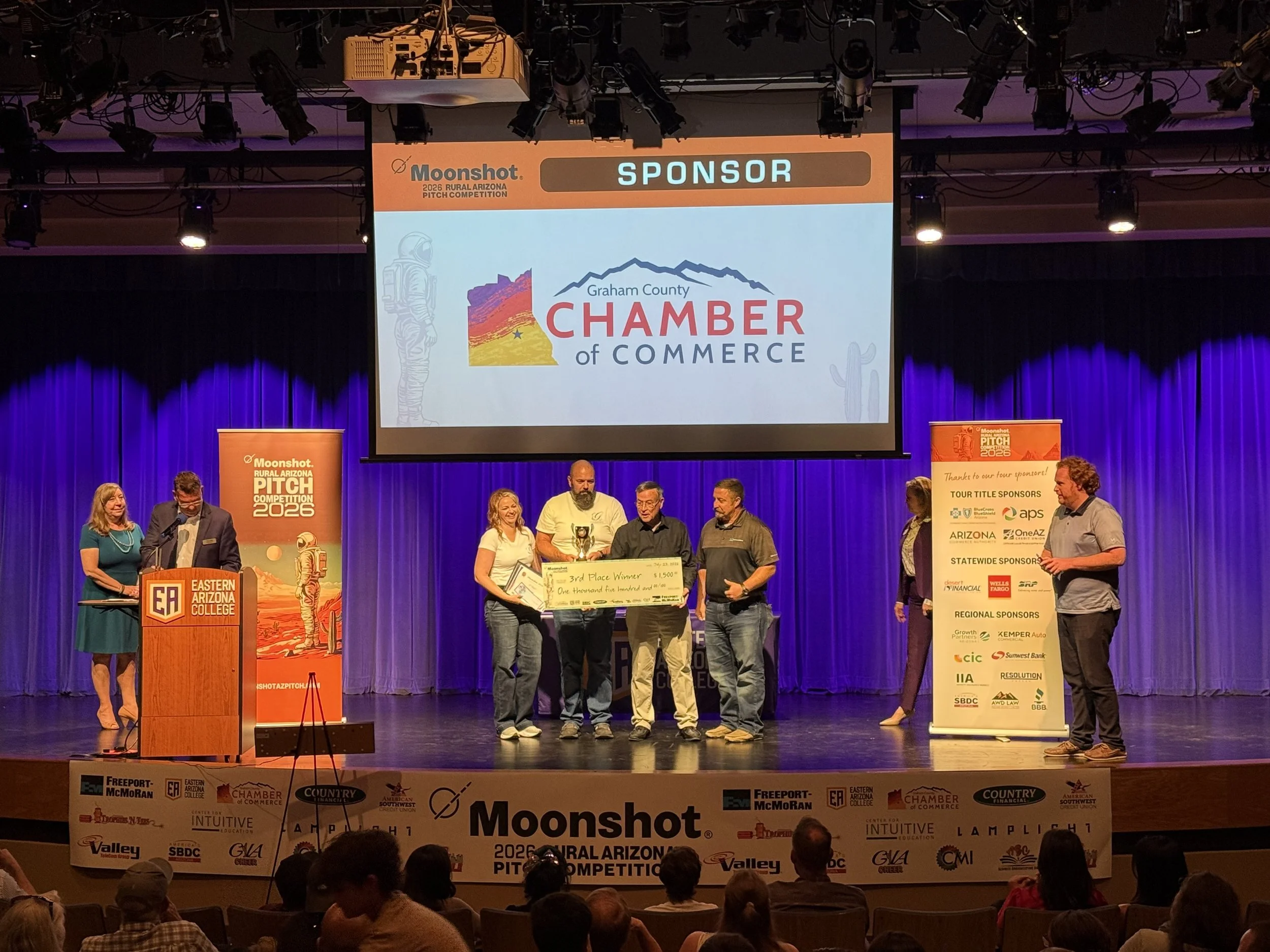 Three people are standing in front of a presentation screen holding a large check for $750, made out to Oli Cakes for third place in the 2025 Rural Arizona Pitch Competition, with sponsors' logos and event details visible on the screen.