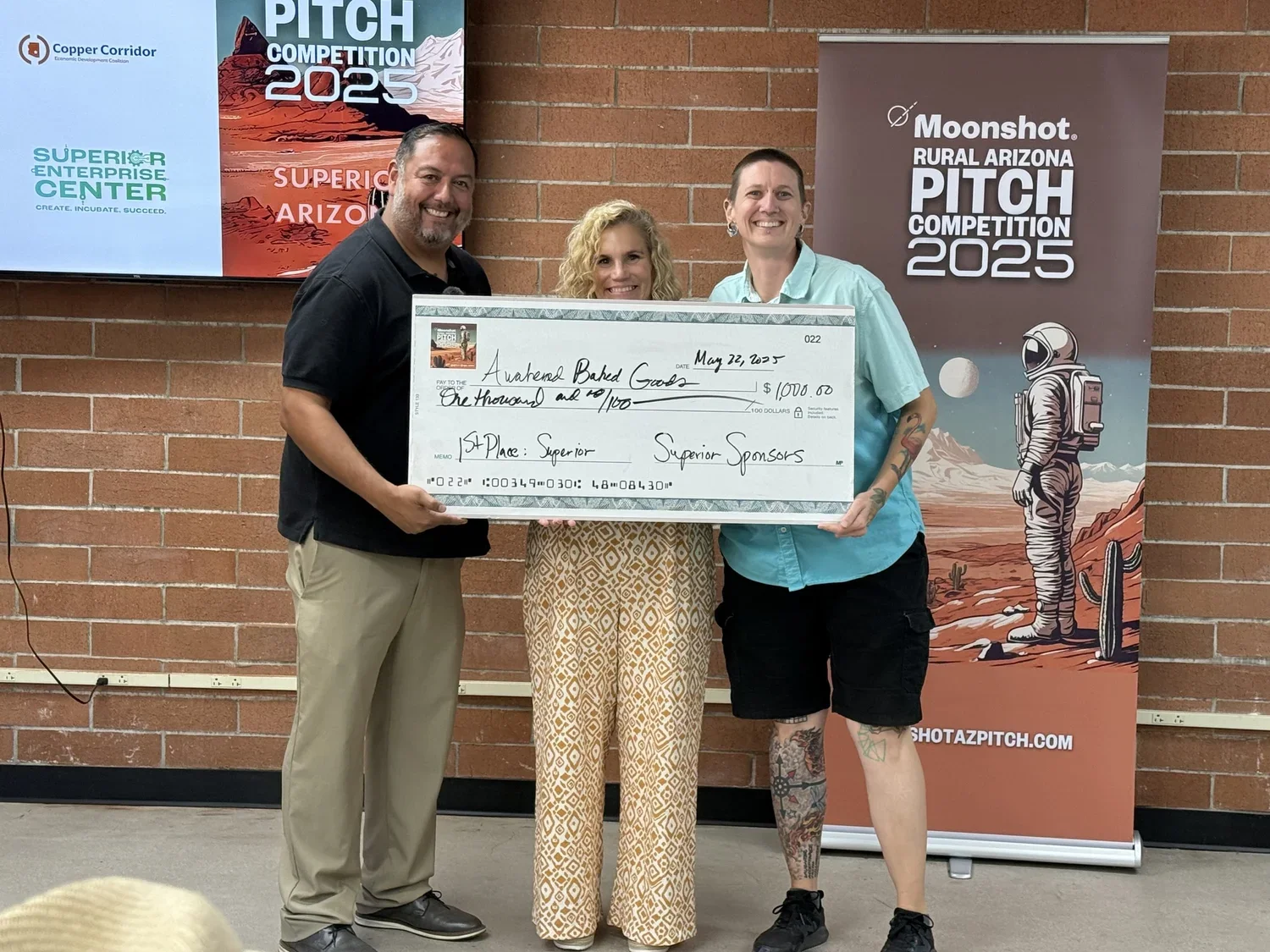 Three individuals on a stage holding a large ceremonial check for $1,000, with two banners in the background. The event appears to be a pitch competition at the Superior Enterprise Center in Arizona.