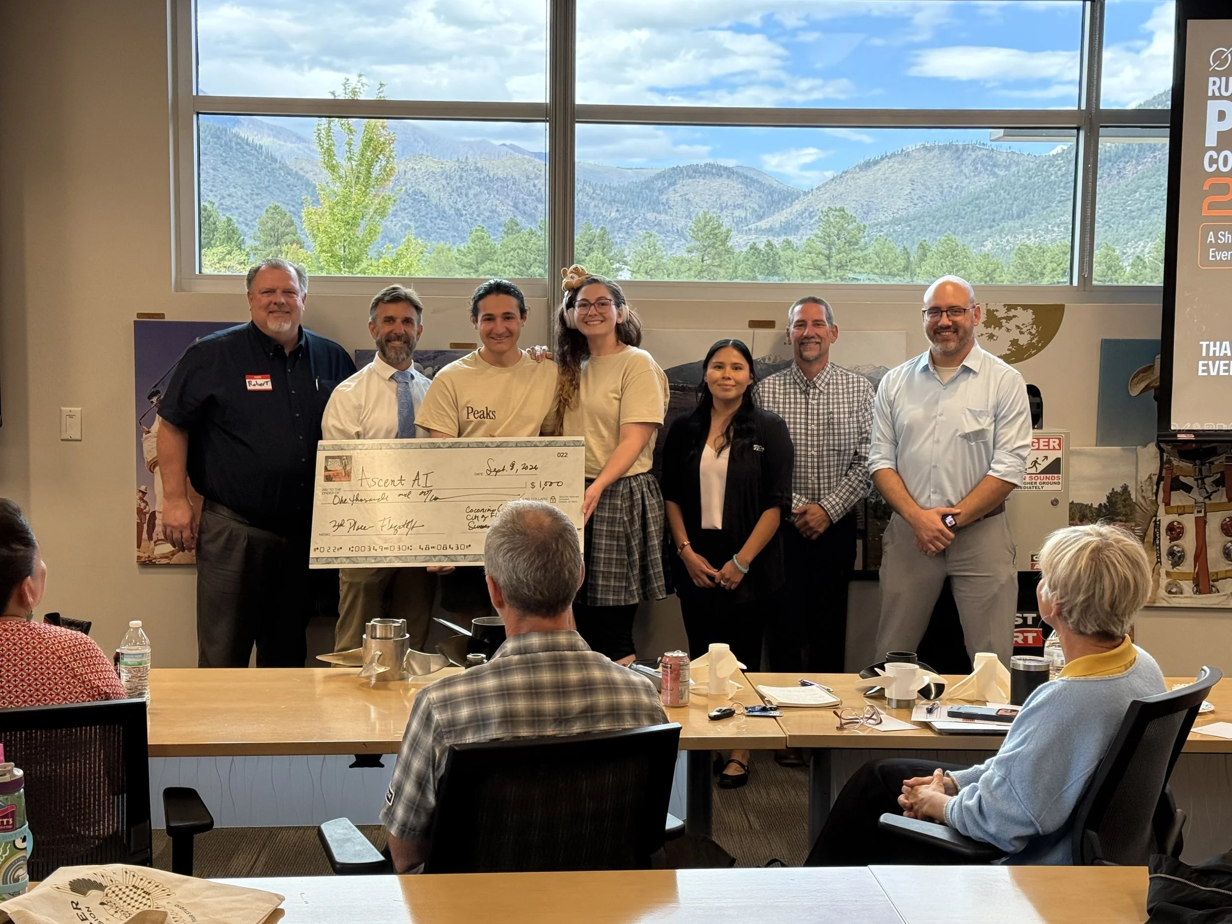 Group of five people standing on a stage, holding a large ceremonial check during the Moonshot Rural Arizona Pitch Competition 2025 event. There are five men and one woman, with a presentation screen behind them displaying event and sponsor logos.