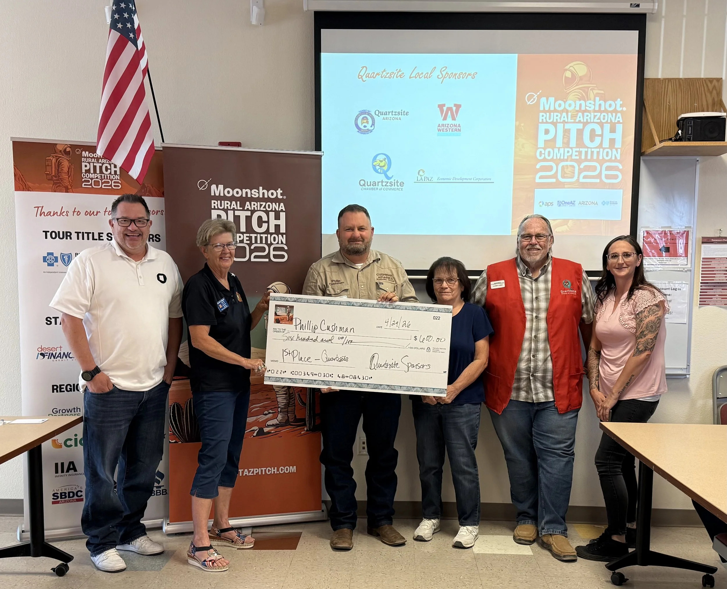 Three people smiling and holding a large novelty check in a room with a presentation setup and signage for the Moonshot Rural Arizona Pitch Competition 2025. An American flag is visible in the background.