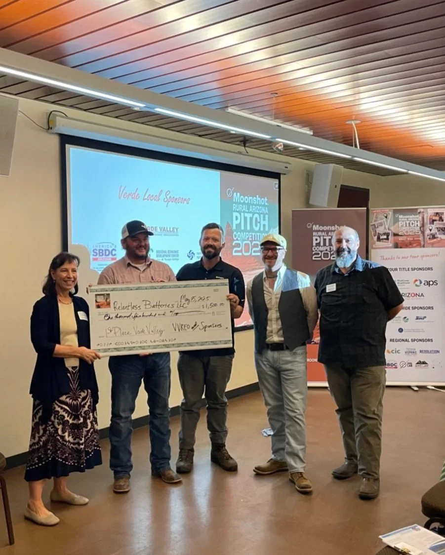 Group of five people standing indoors, holding a large check at an award ceremony, with a presentation screen in the background displaying logos and event details.
