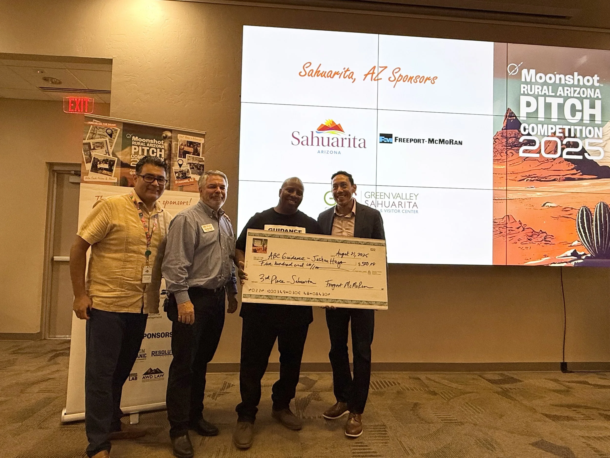 Four men standing on a stage with a large check, celebrating at the Moonshot Rural Arizona Pitch Competition 2025 in Sahuarita, Arizona. Behind them is a large screen displaying event logos and sponsors.