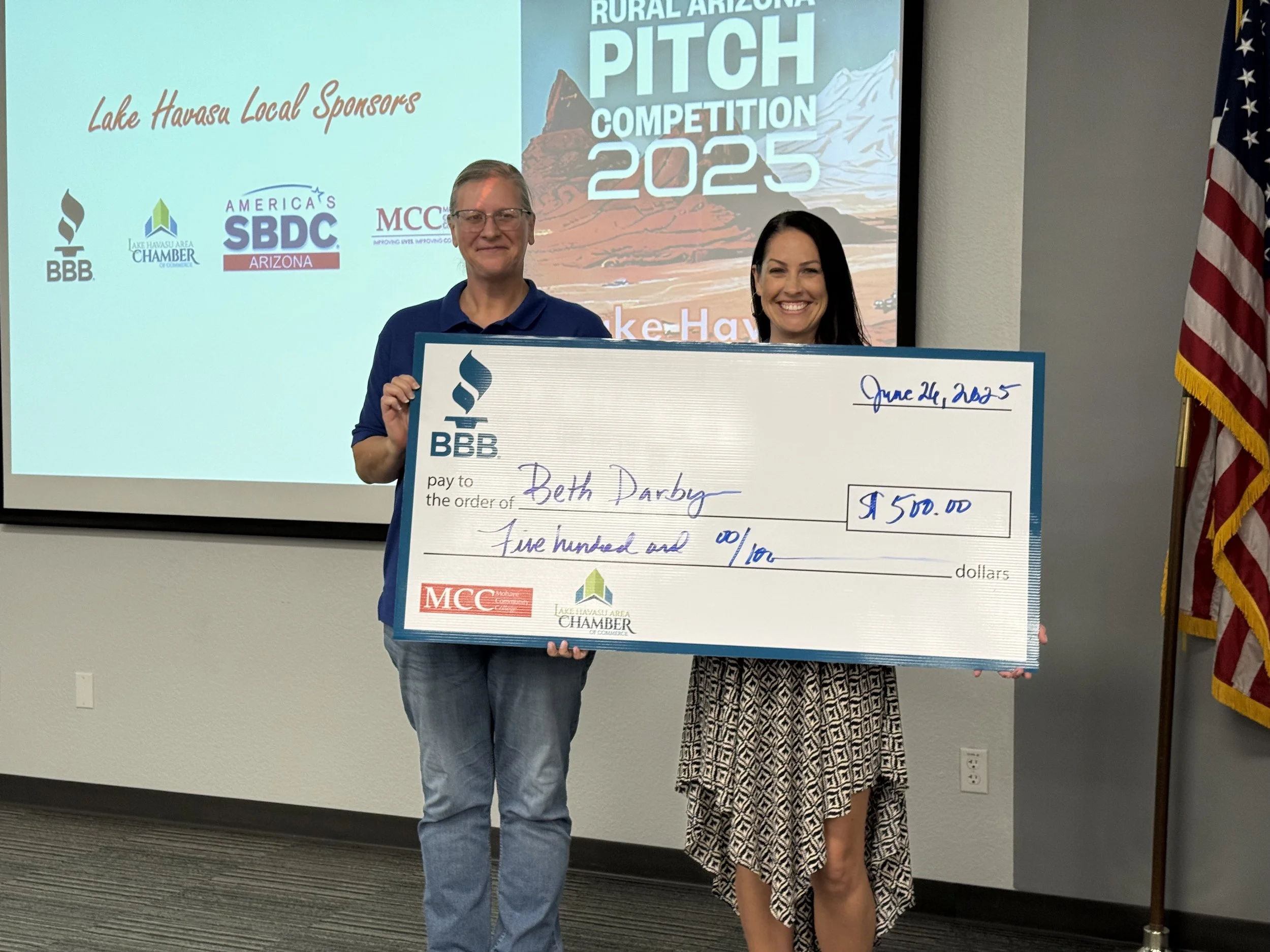 Two women holding a large ceremonial check for $500, standing in front of a presentation screen at an event. The screen displays logos of local sponsors for the Lake Havasu Rural Arizona Pitch Competition 2025. One woman is wearing glasses and a blue shirt, the other is smiling in a patterned dress.