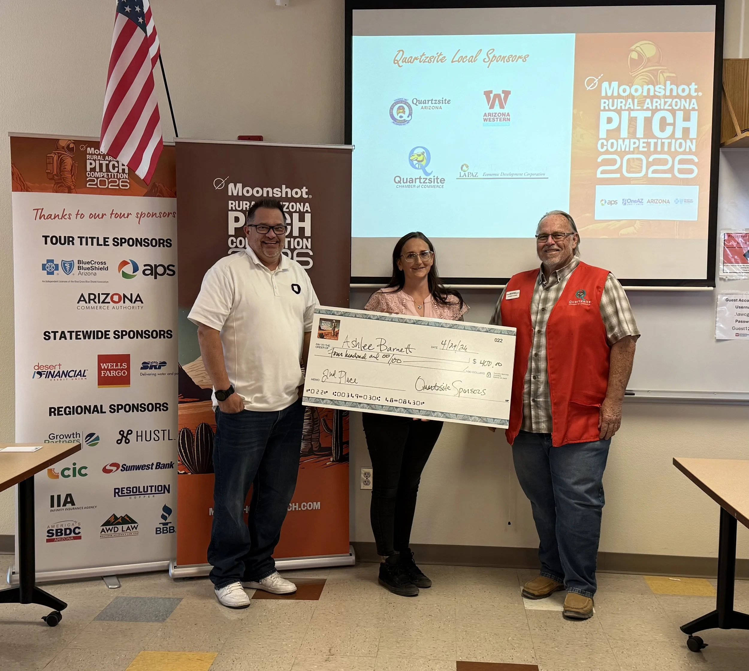 Three men holding a large ceremonial check at the Moonshot Rural Arizona Pitch Competition event. An American flag is visible in the background along with event banners.