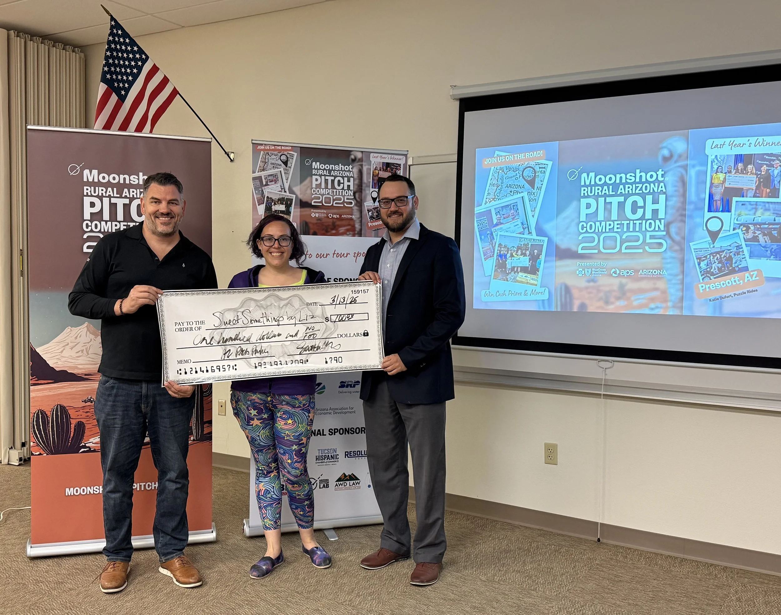 Three people holding a large check in front of a "Moonshot Rural Arizona Pitch Competition 2025" banner and screen. An American flag and posters are displayed in the background.