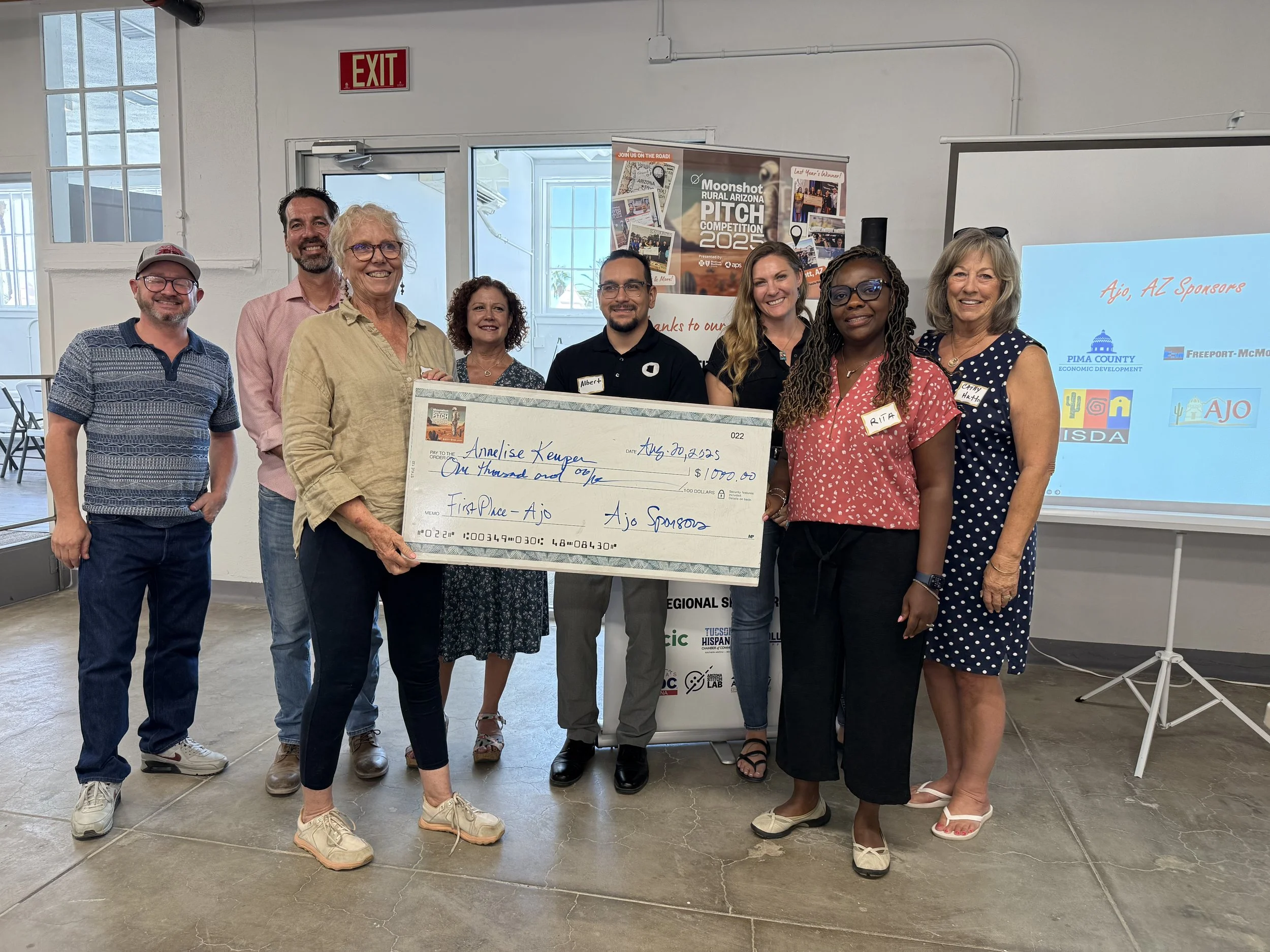 Group of nine people standing indoors, holding a large check, at an event for a pitch competition. They are smiling and dressed casually, with a presentation screen and posters in the background.