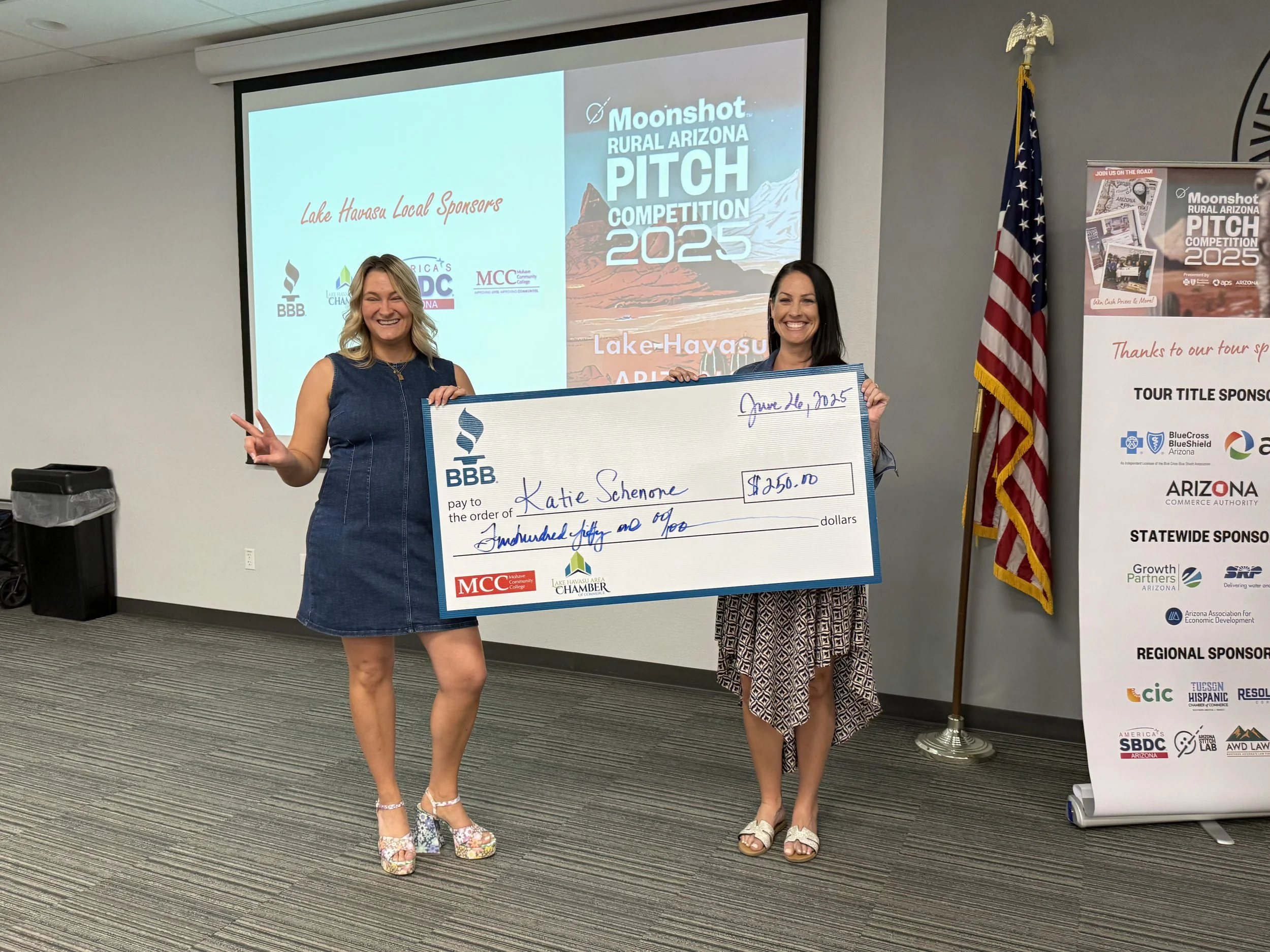 Two women smiling and holding a large check at an indoor event, with a presentation screen displaying 'Moonshot Rural Arizona Pitch Competition 2025' behind them. One woman is making a peace sign. The check is for $250.00 made out to Katie Scherne. An American flag is standing nearby.