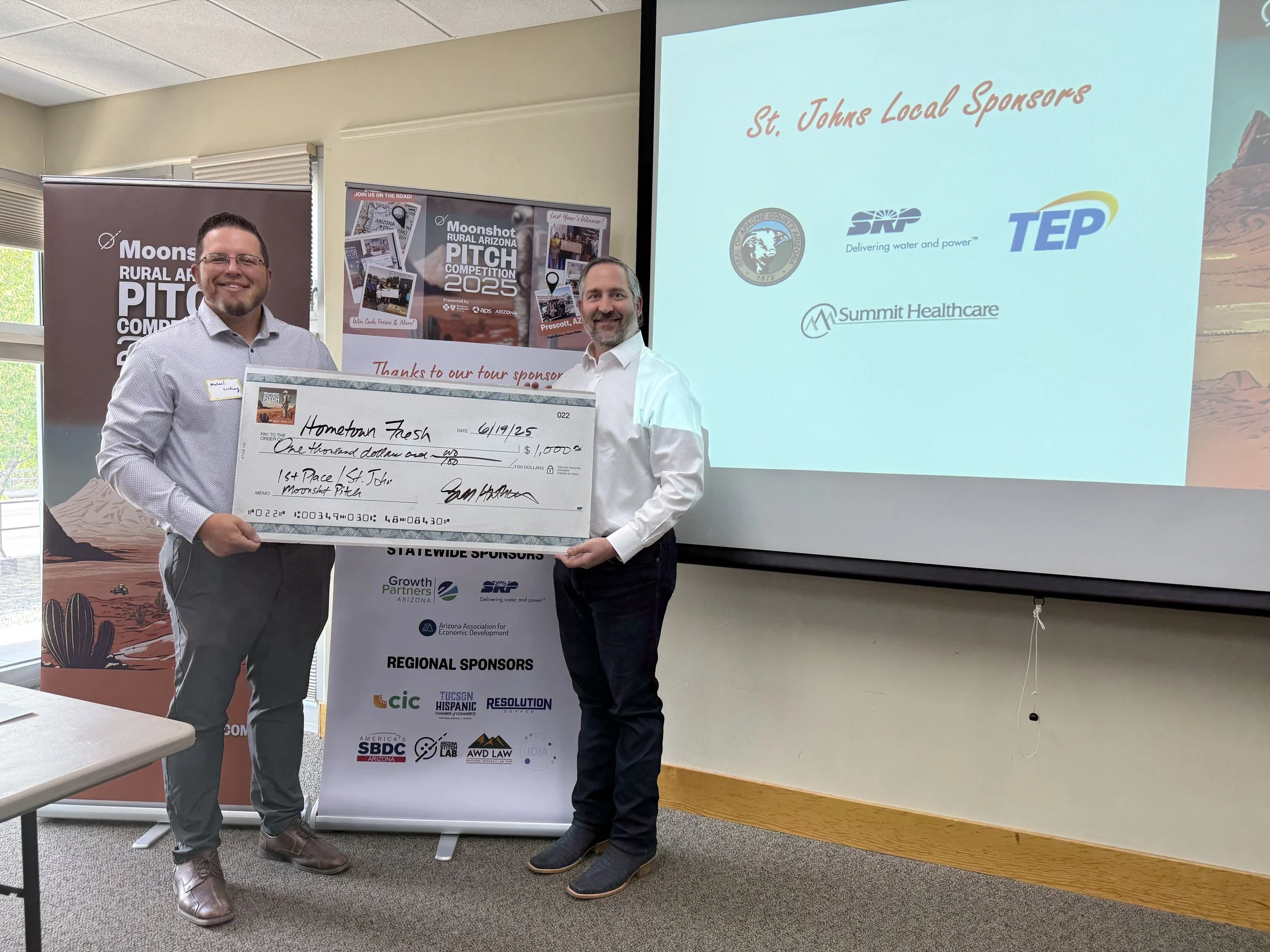 Two men standing in a room holding an oversized check for $1,000, with a presentation screen behind them displaying logo sponsors, and a banner with event details. The man on the left is smiling, wearing glasses, a light-colored shirt, and dark pants. The man on the right is smiling, wearing a white shirt and dark pants.