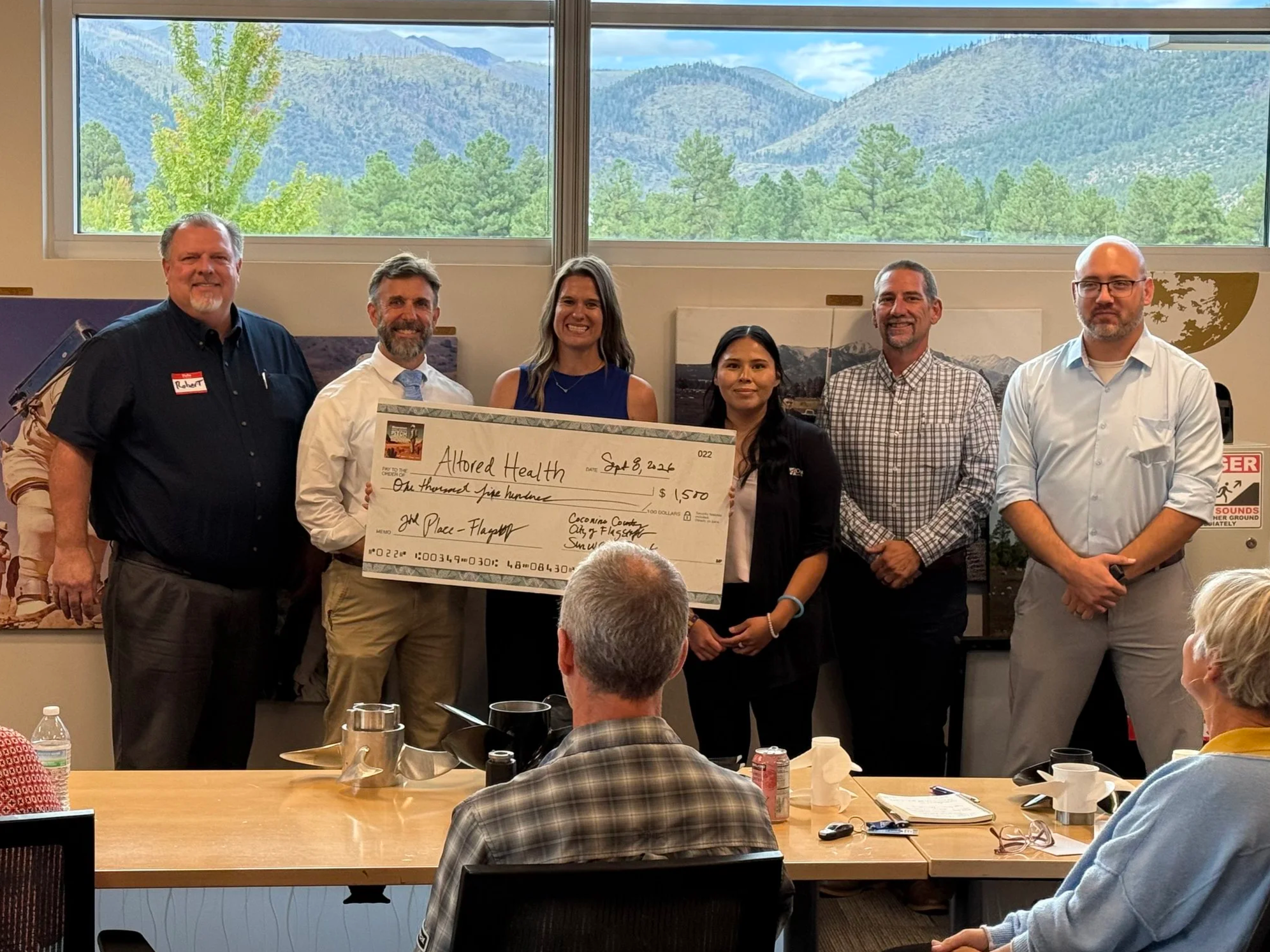Group of five people standing together indoors, holding a large ceremonial check, with a presentation screen in the background displaying event details and sponsor logos.
