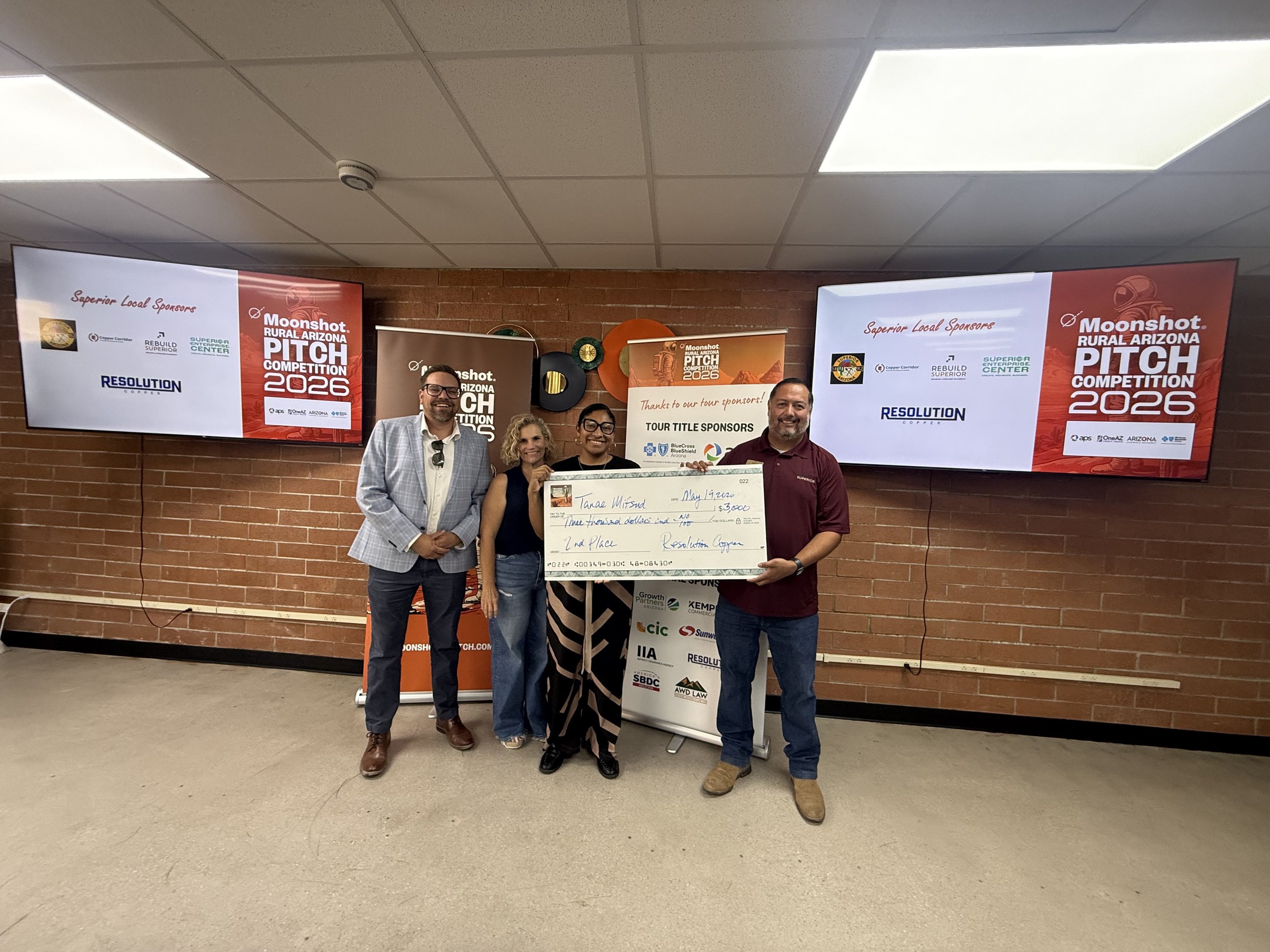 Three people standing indoors holding a large check for $500, posing for a photo during the Moonshot Rural Arizona Pitch Competition 2025. The man on the left wears a black shirt and beige pants, the woman in the middle has blonde curly hair and patterned pants, and the woman on the right wears glasses, a burnt orange shirt, and jeans. A large screen and a poster with space-themed desert artwork are visible in the background.