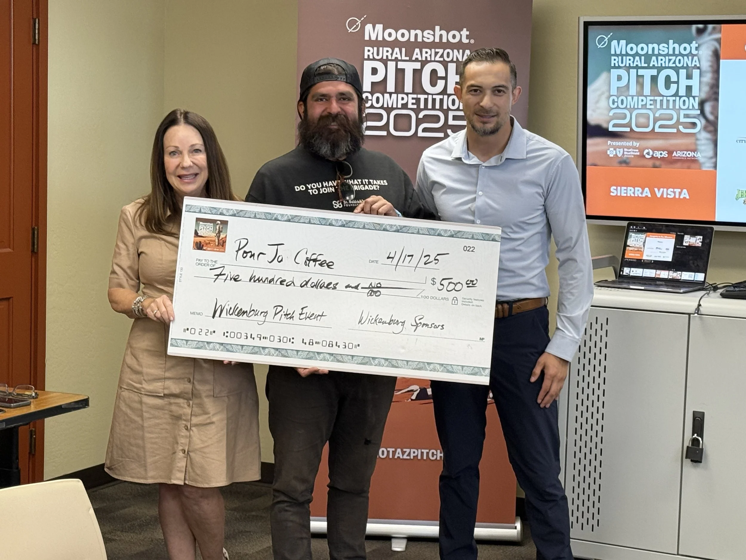 Three people holding a large check at the Moonshot Rural Arizona Pitch Competition 2025. The check is made out to "Pour Jo Coffee" for $500, dated 4/17/25. Signage in the background shows the event logo and details.