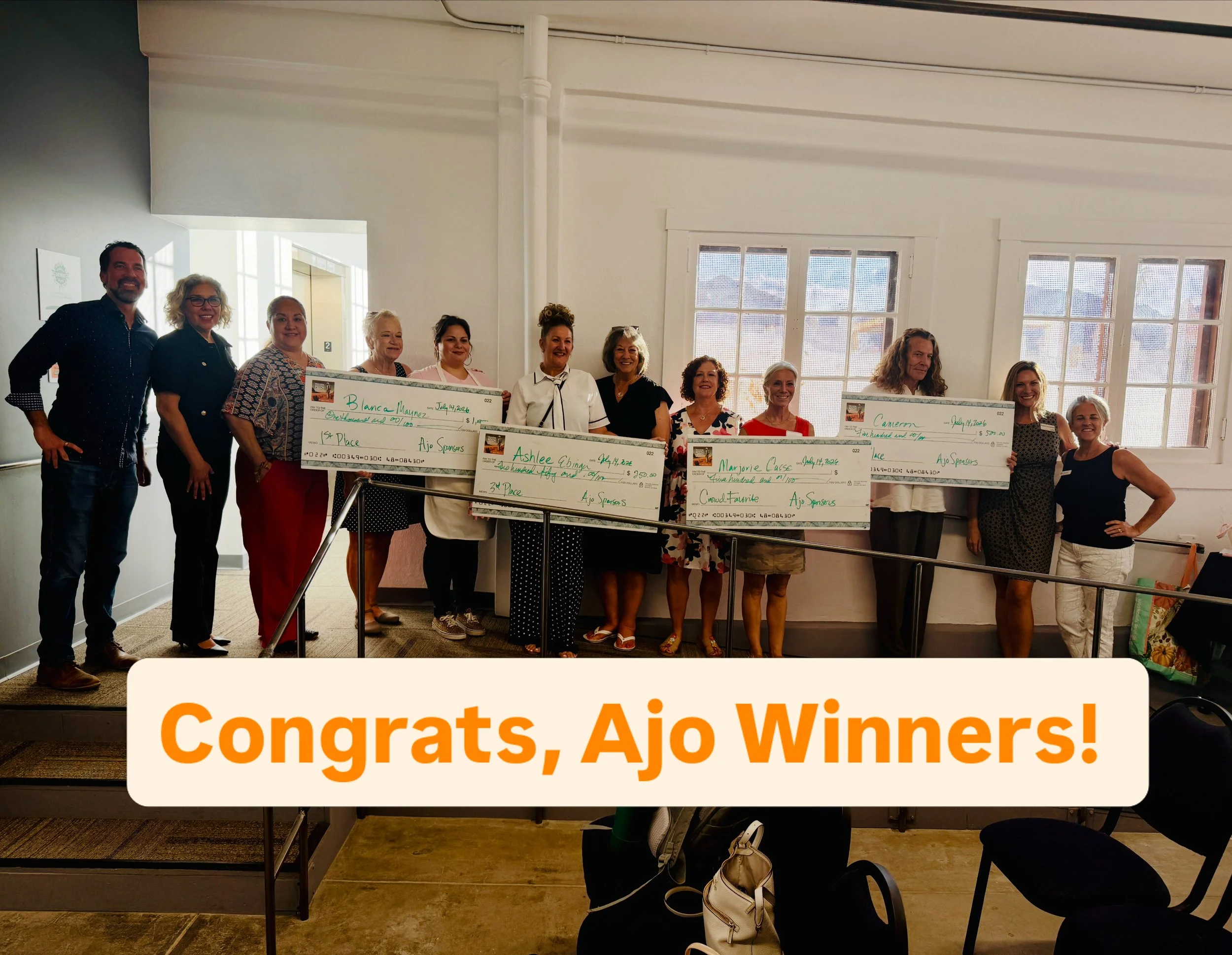 Group of nine people standing indoors, holding a large check, at an event for a pitch competition. They are smiling and dressed casually, with a presentation screen and posters in the background.