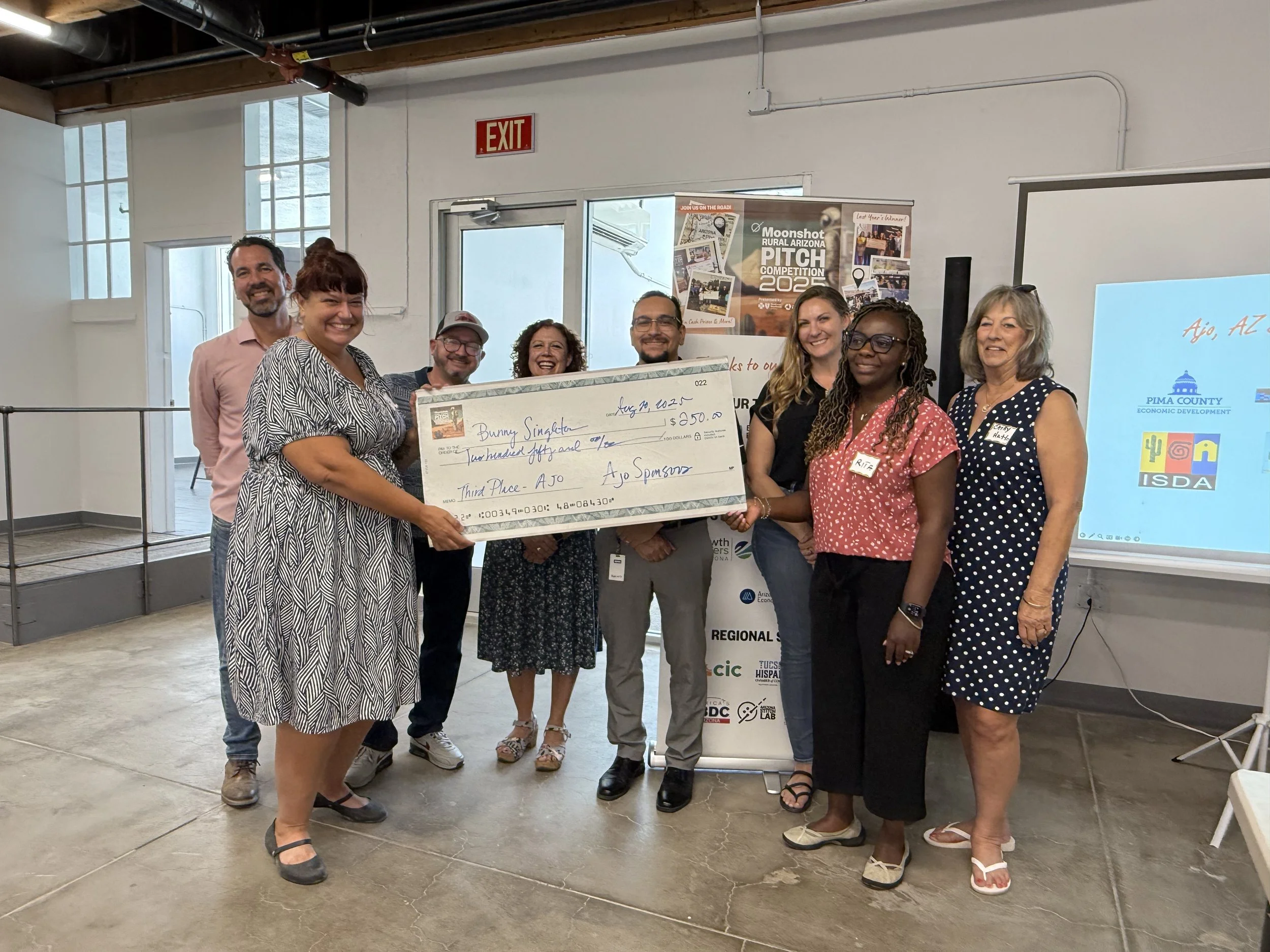Group of eight people standing indoors, holding a large check awarded for third place at the Moonshot Rural Arizona Pitch Competition 2022. The check is made out to Bunny Sinleton for $250. All are smiling, some wearing name tags, with a screen and a banner in the background.