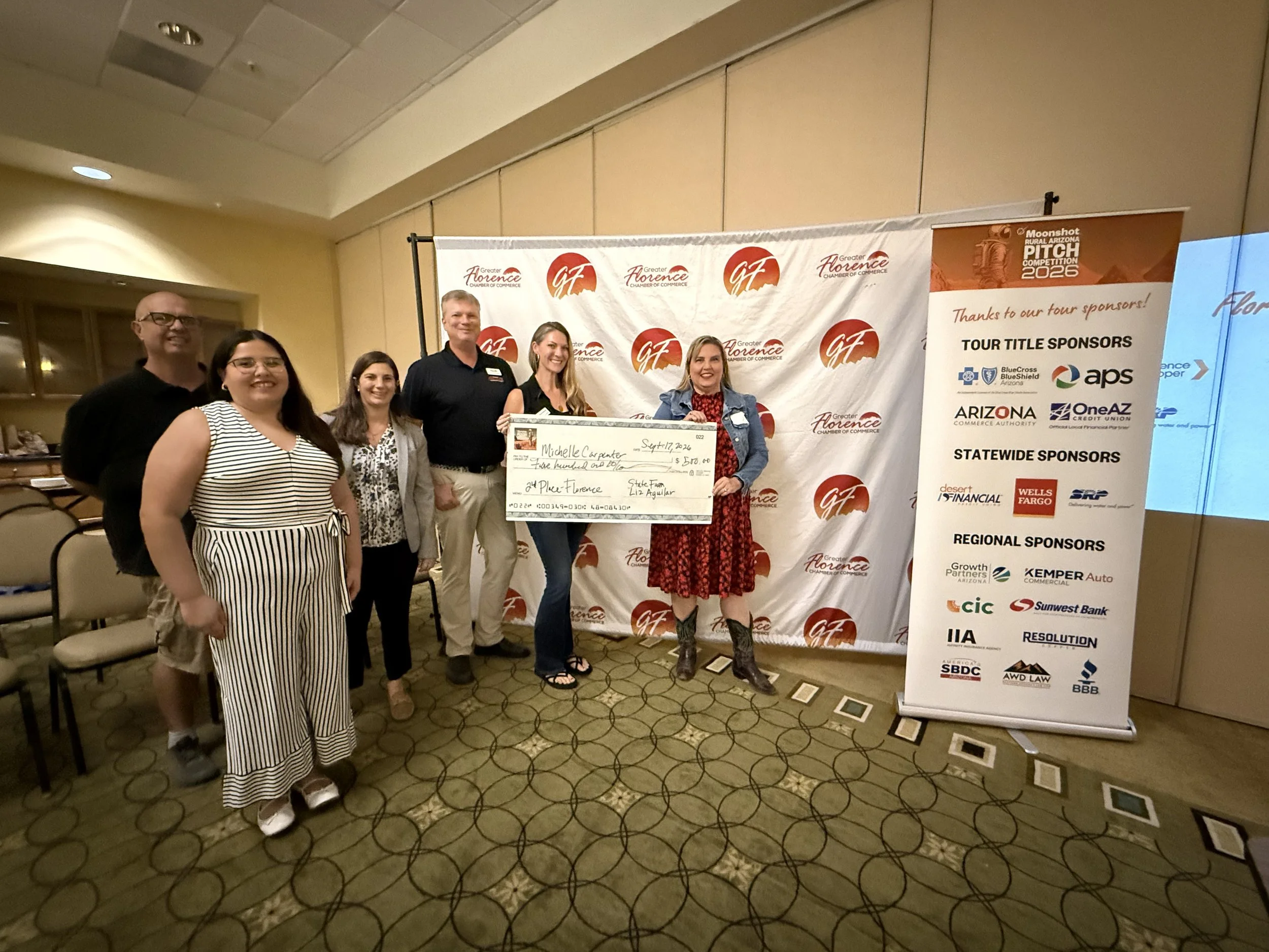 Group of five people posing indoors at a chamber of commerce event, holding a large check, with a backdrop displaying sponsors' logos and banners for the event.