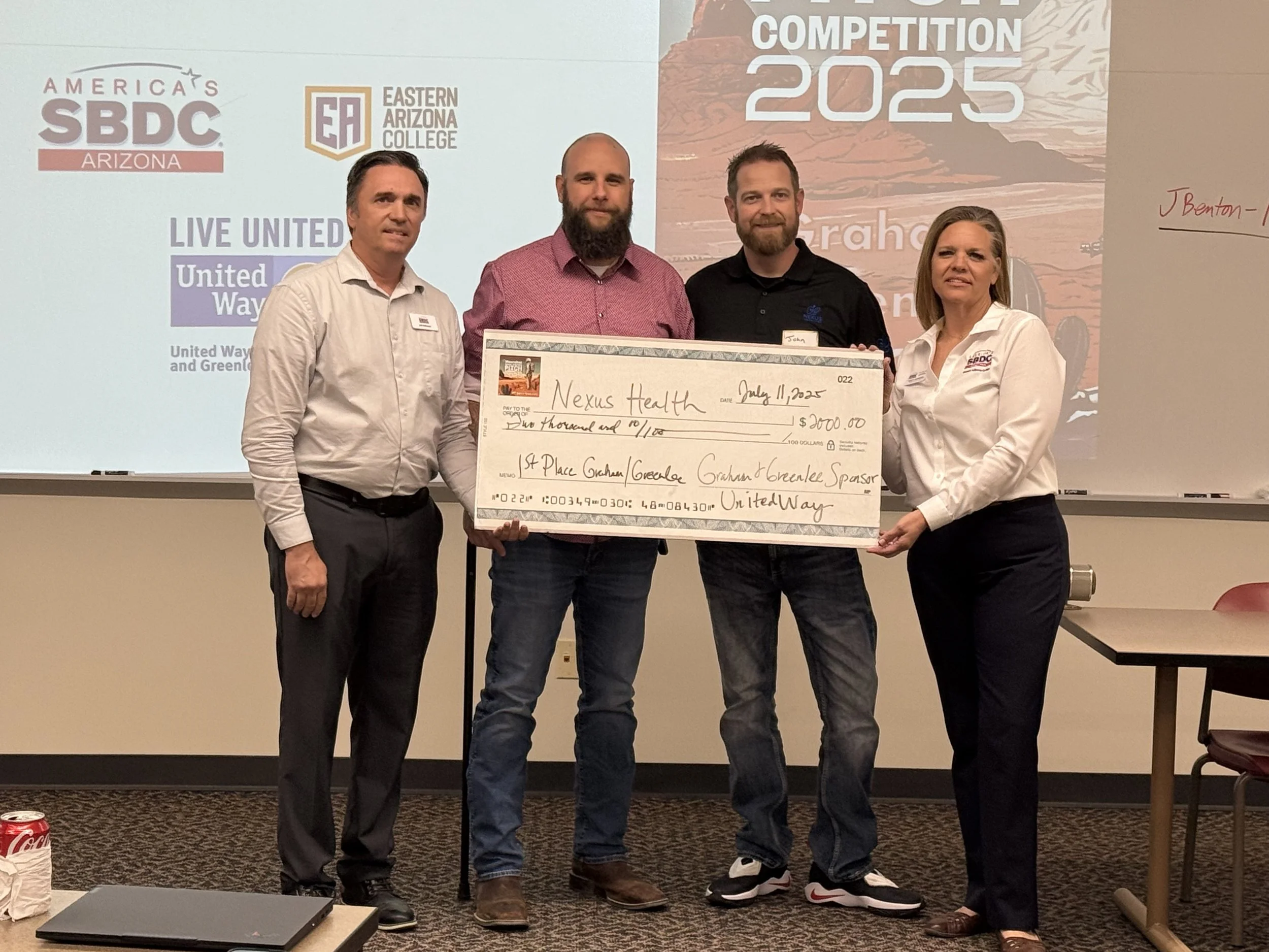 Four people standing in a room, holding an oversized check made out to Nexus Health for $2,000, during an award or donation event, with a presentation screen in the background displaying logos of America’s SBDC Arizona, Eastern Arizona College, and the United Way.