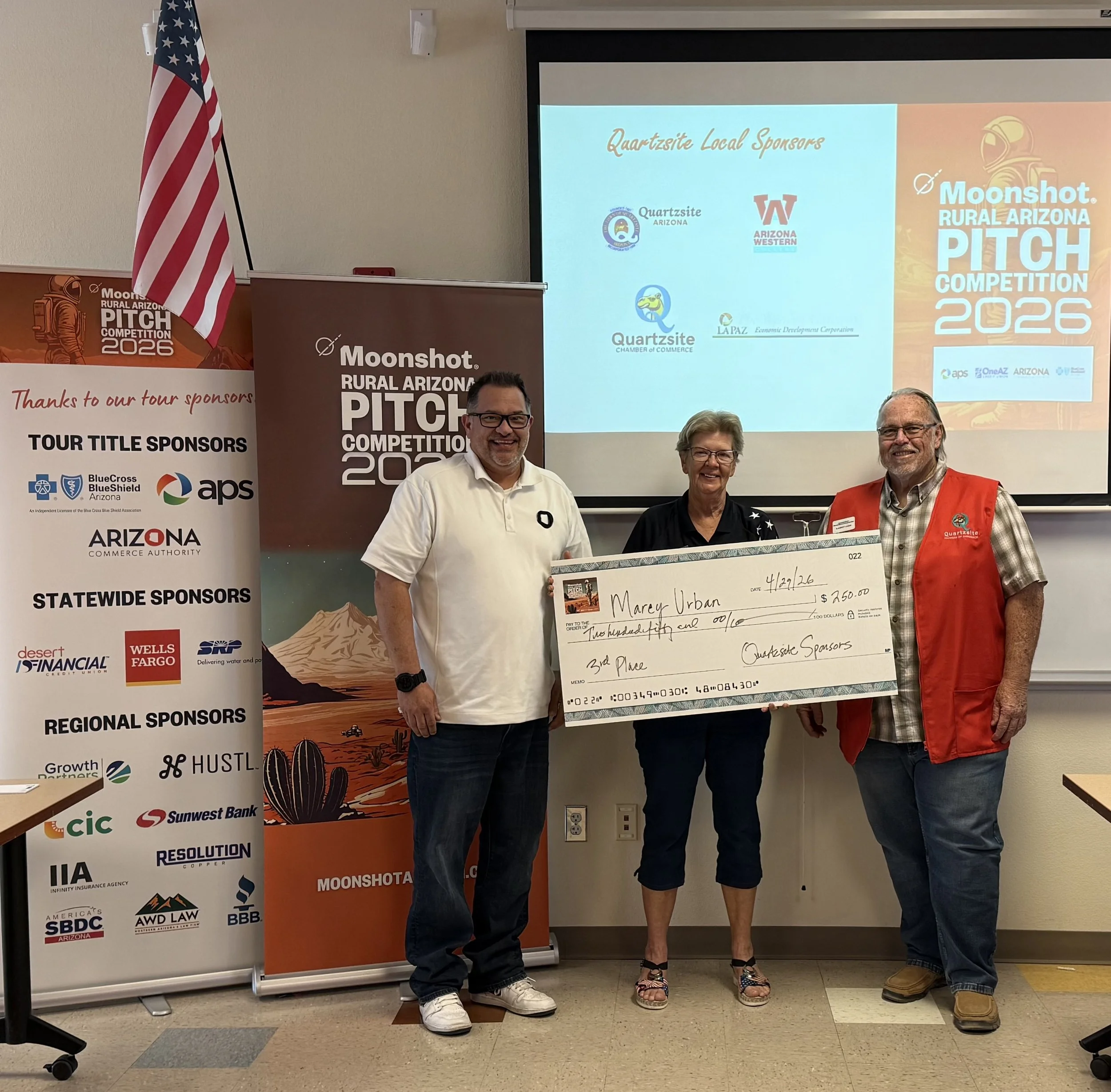 Two people holding a large check in front of a banner for Moonshot Rural Arizona Pitch Competition 2023. The check displays an amount of $200 commended to "Whole Bed Prep Clinic." There is an American flag and sponsor logos in the background.