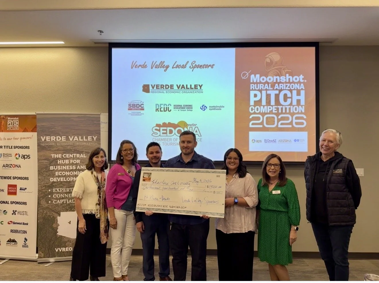 Group of five people holding an oversized check at an event, possibly a competition or business pitch, with a projection screen and banners in the background. The check is awarded to 'Relentless Batteries LLC' for $1,500. The event is titled 'Moonshot Rural Arizona Pitch Competition 2023.'