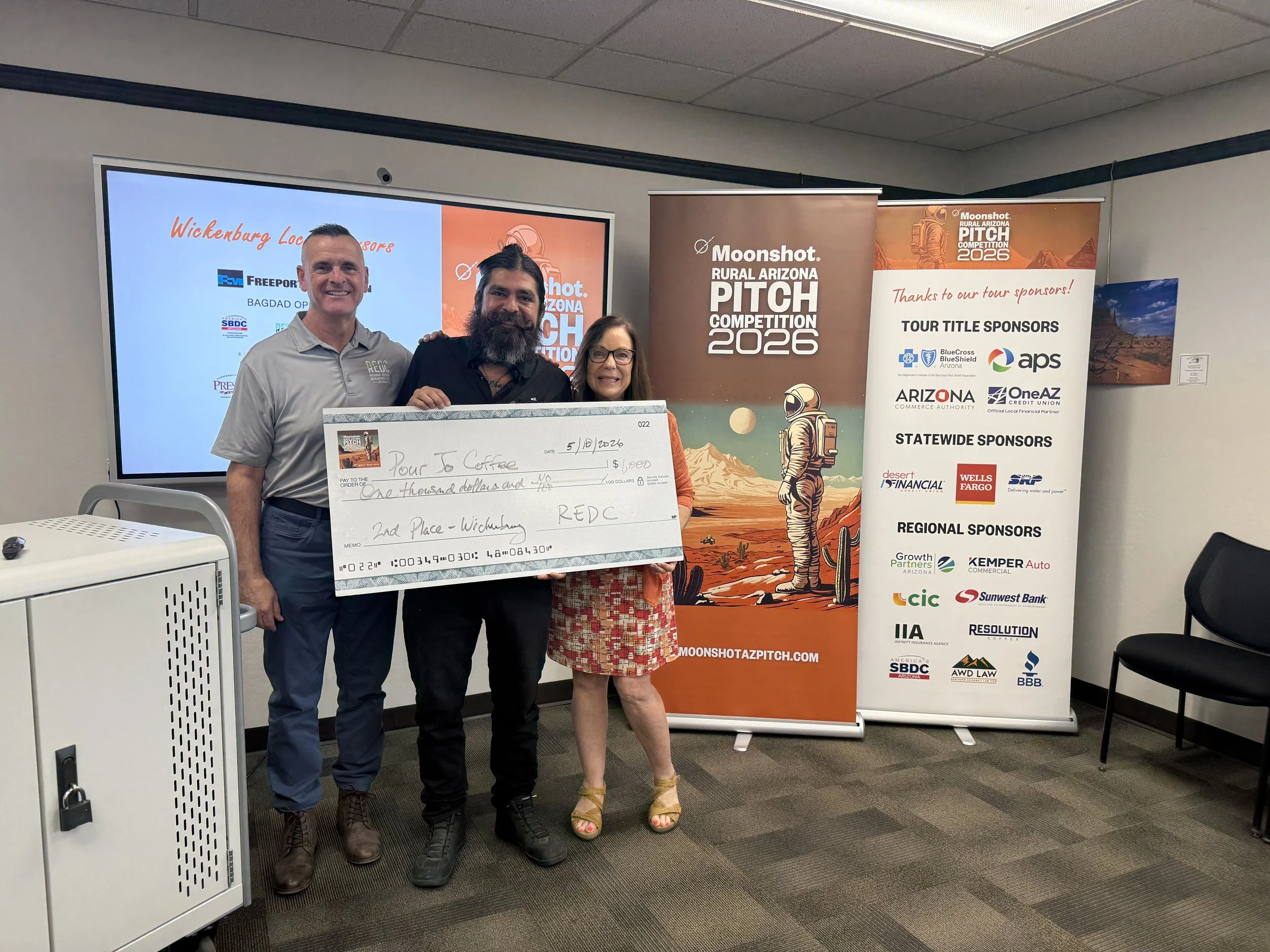 Three people holding a large check at the Moonshot Rural Arizona Pitch Competition 2025. The check is made out to "Pour Jo Coffee" for $500, dated 4/17/25. Signage in the background shows the event logo and details.