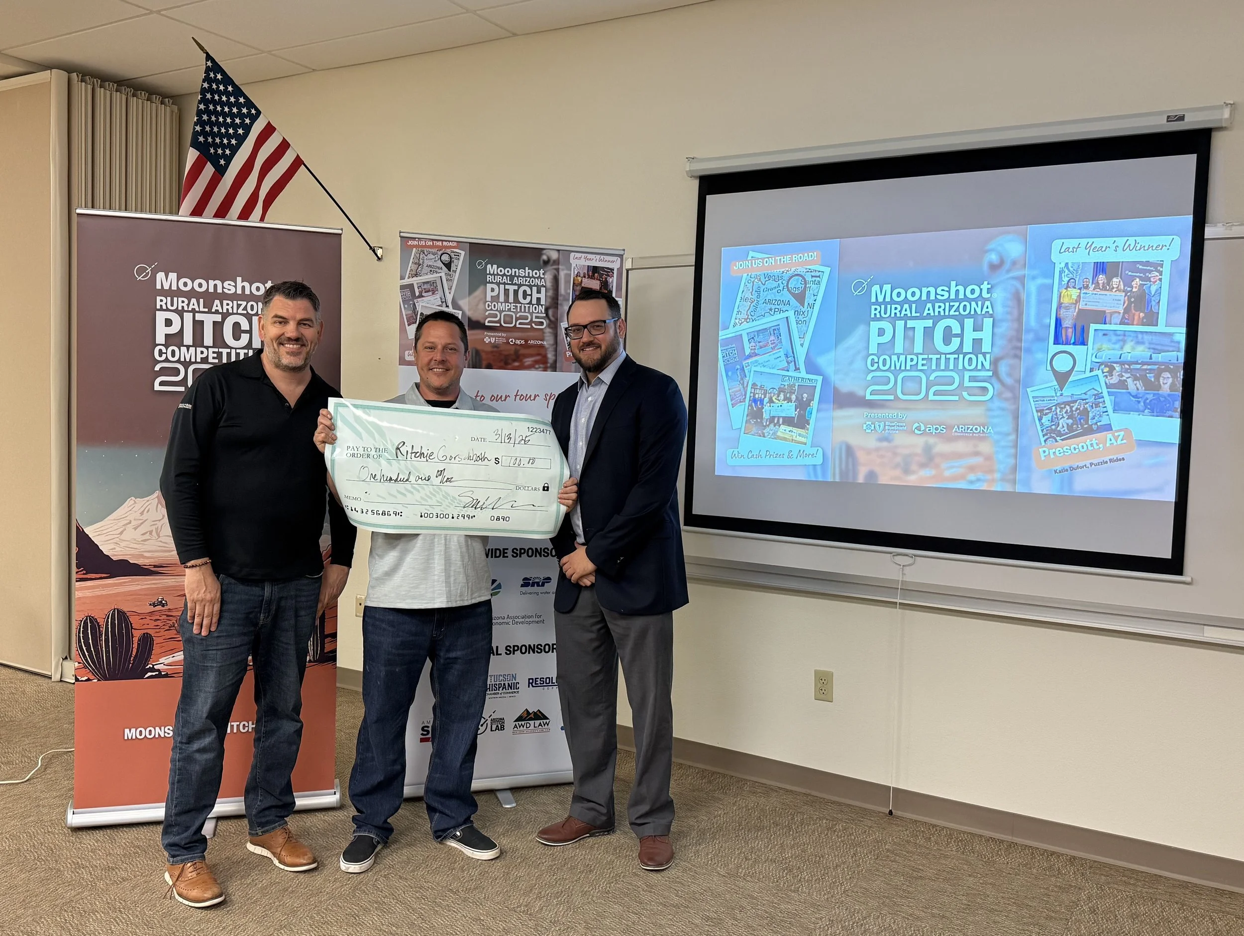Three men standing in front of Moonshot Rural Arizona Pitch Competition 2025 banners, one holding a large check. An American flag is on the wall.
