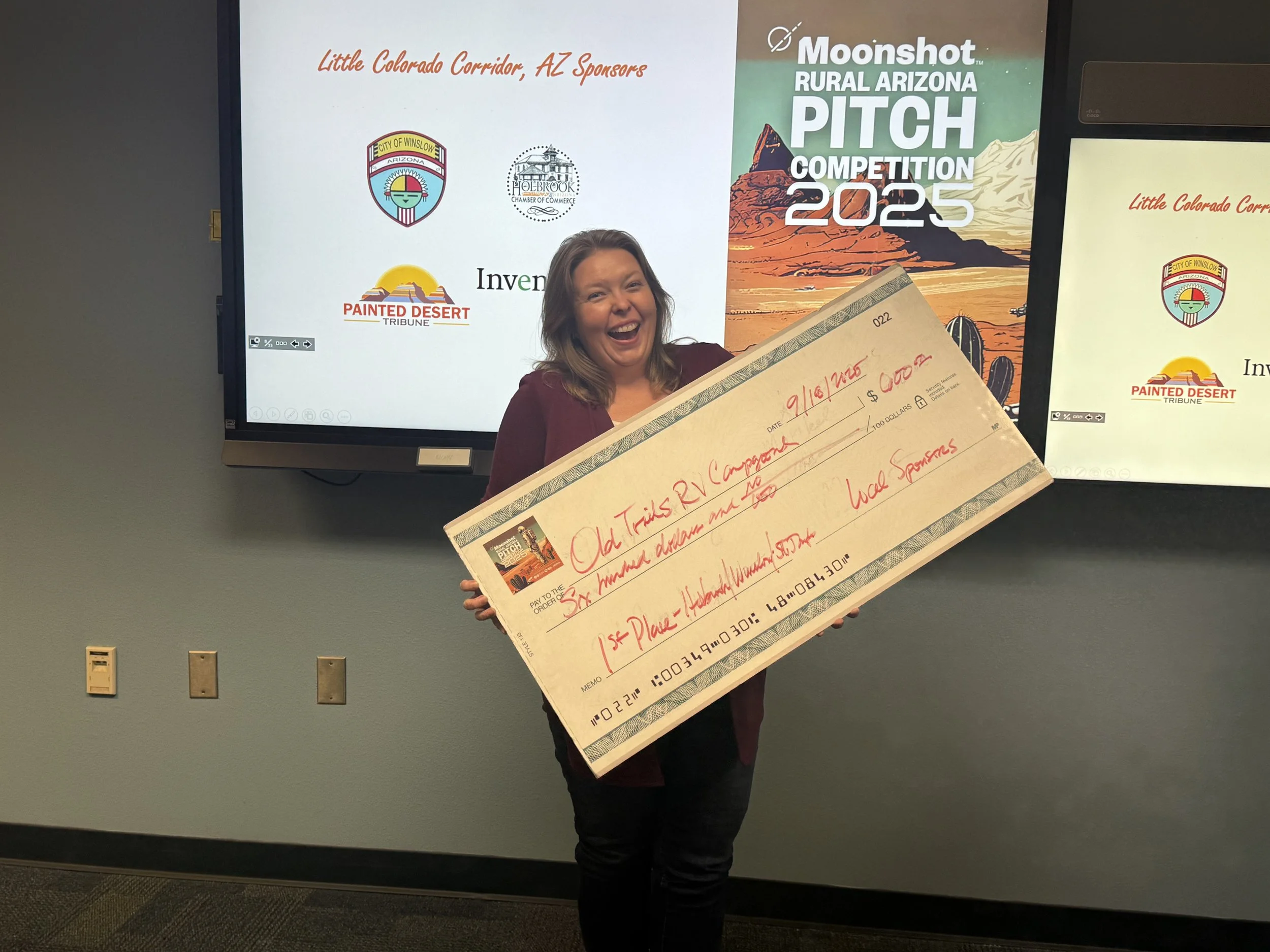 A woman smiling and holding a large check in front of two screens displaying the 'Moonshot Rural Arizona Pitch Competition 2025' logo and other sponsors, celebrating her award at the event.