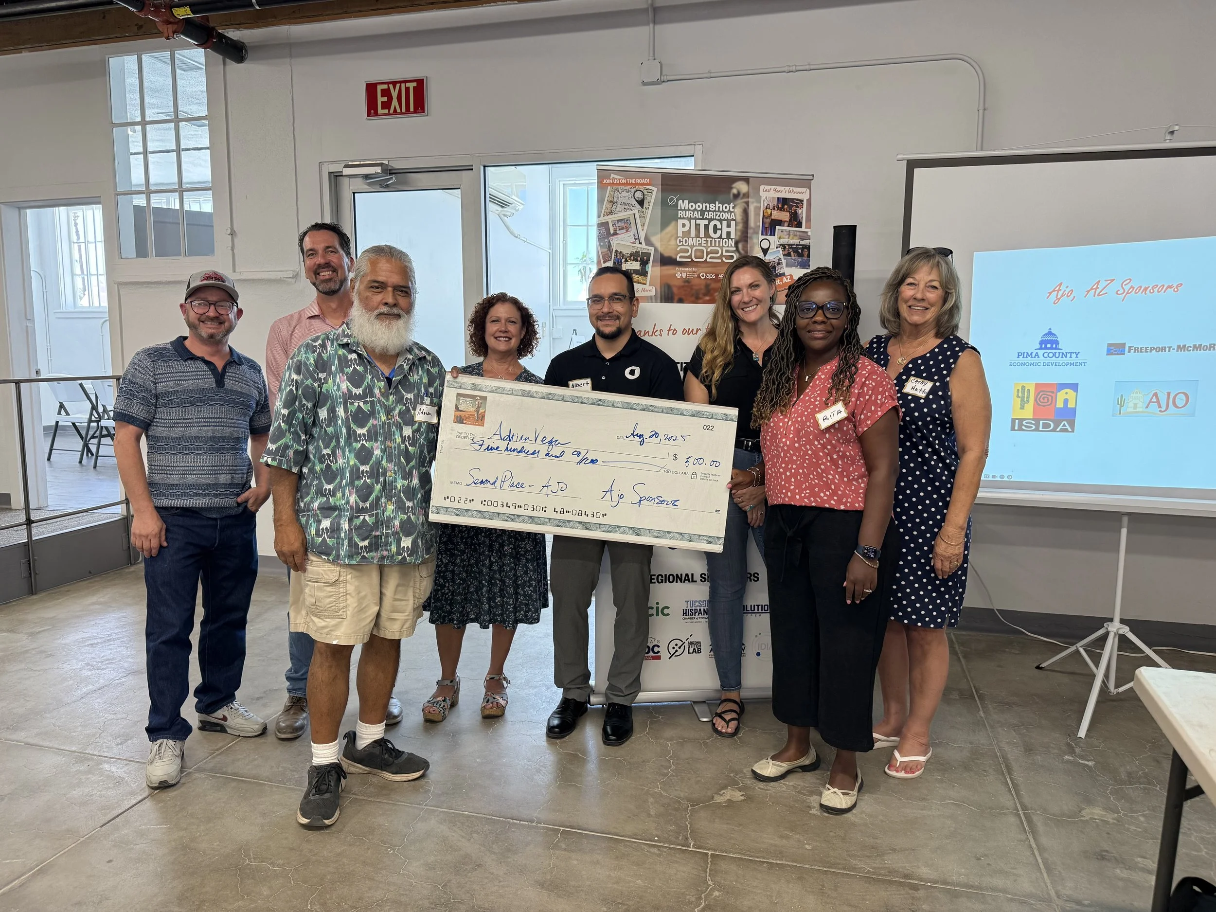 Group of nine people standing indoors, holding a large check for $600, at the Moonshot Rural Arizona Pitch Competition 2025. The group includes diverse men and women, some wearing name tags, with a projector screen and a poster in the background.