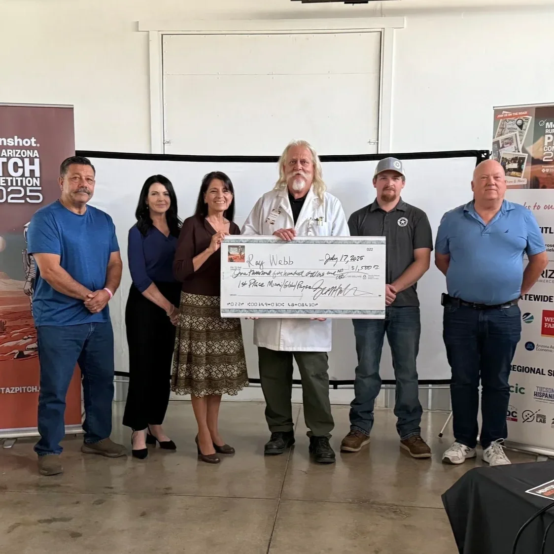 Group of six people standing together, holding a large ceremonial check, at an indoor event with banners in the background.