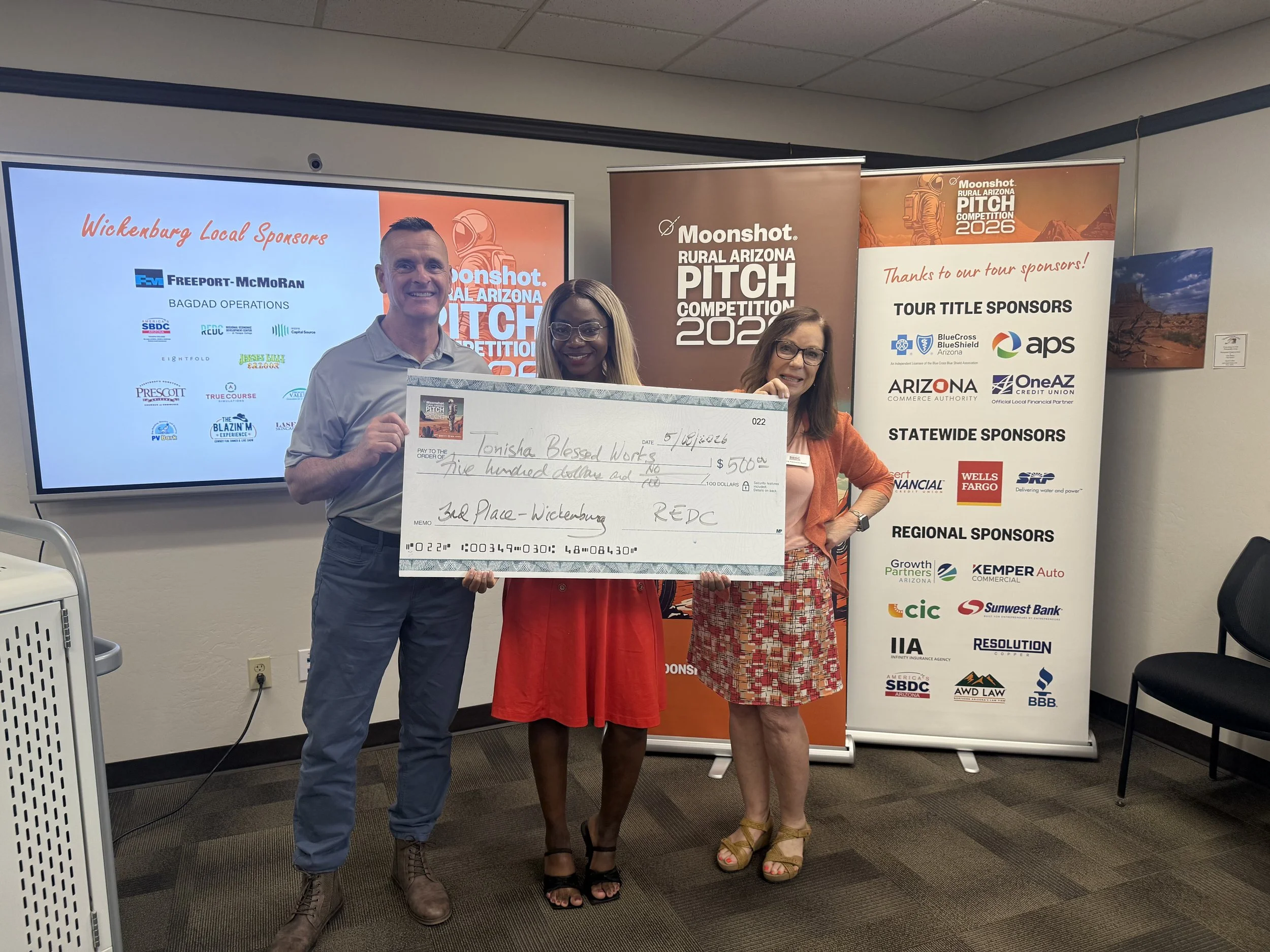 Three people standing indoors at a conference or event, holding a large check, with banners and a screen displaying the event's information in the background. The man on the left has short hair and is wearing a gray shirt, the woman in the middle has glasses and a red dress, and the woman on the right has glasses and a patterned skirt with an orange top.