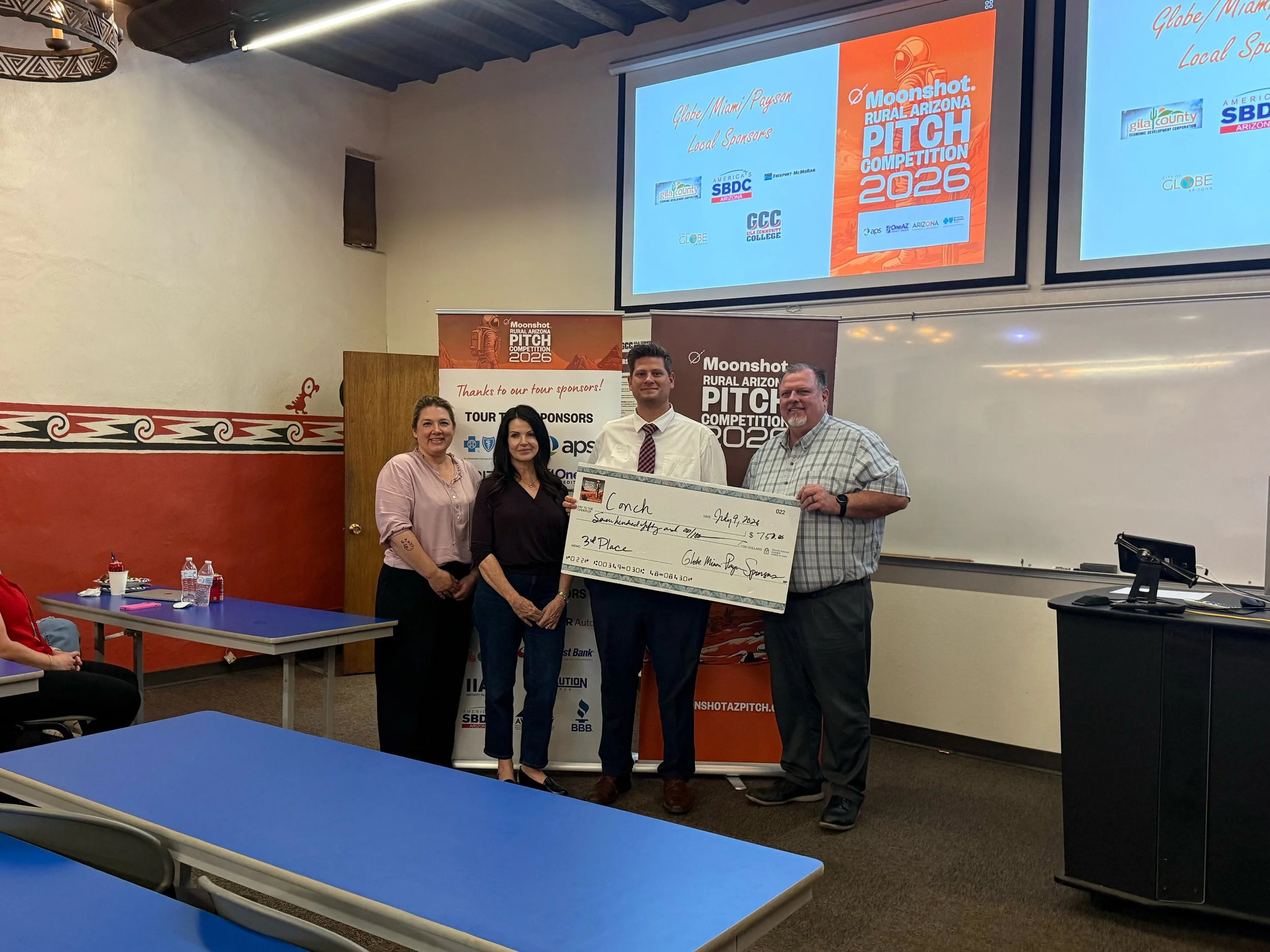 Group of six people standing indoors, holding a large check during a celebratory event for the Moonshot Rural Arizona Pitch Competition 2025. There are promotional banners on each side, and a white wall with air conditioning units above.