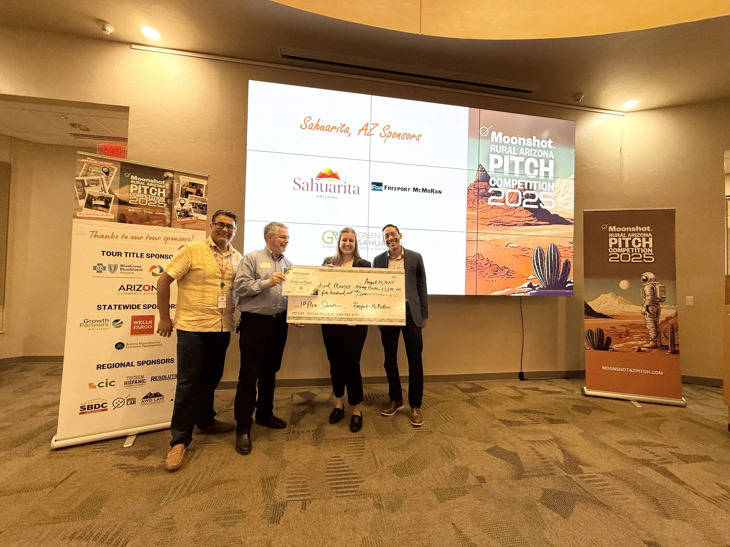 A group of four people standing in front of a large screen and banners at the Moonshot Rural Arizona Pitch Competition 2025; two men and two women, one woman holding a large check.