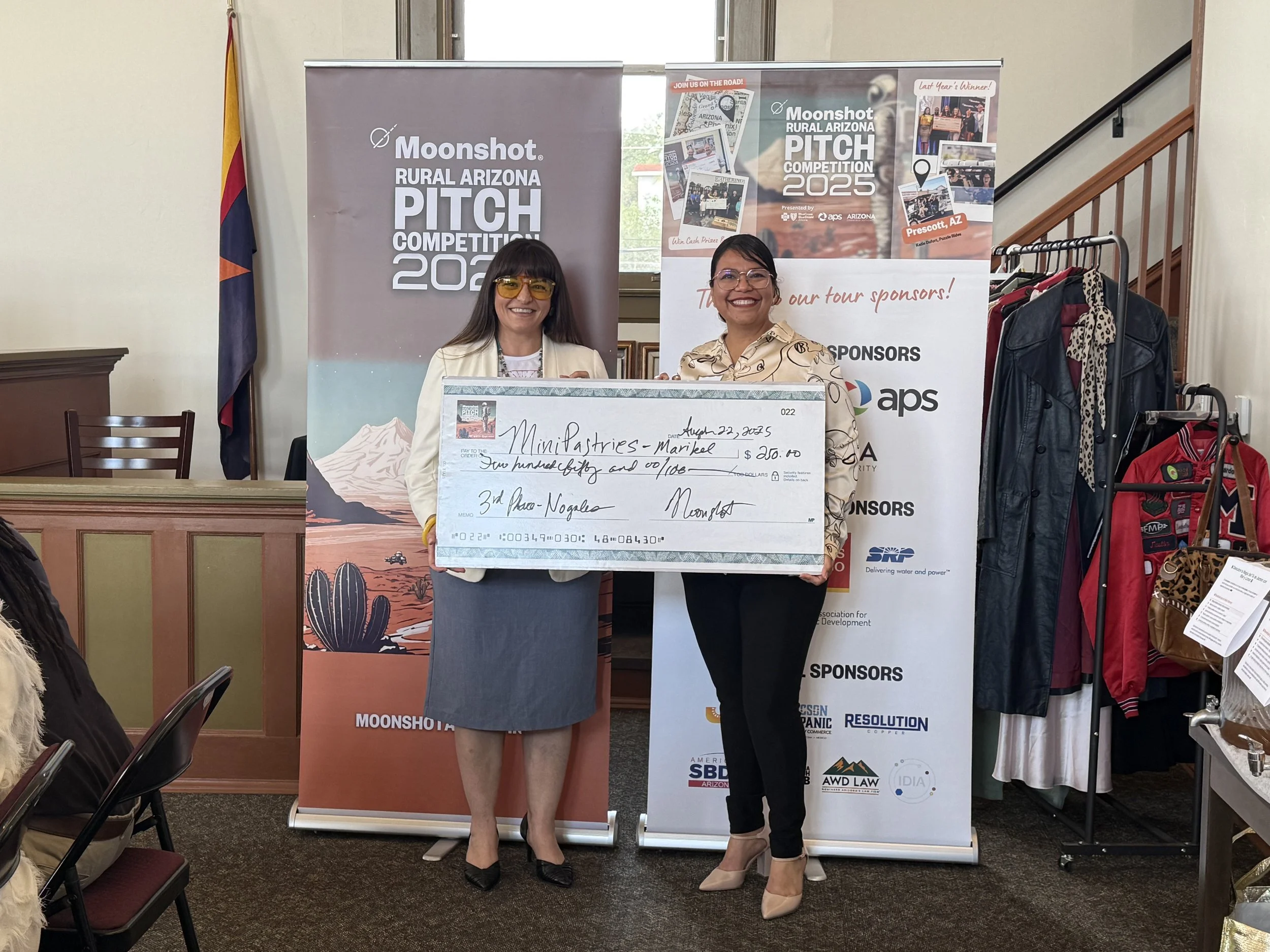 Two women standing indoors holding a large ceremonial check at the Moonshot Rural Arizona Pitch Competition 2023. Behind them are banners and clothing racks, with one banner displaying the event's logo and sponsors.