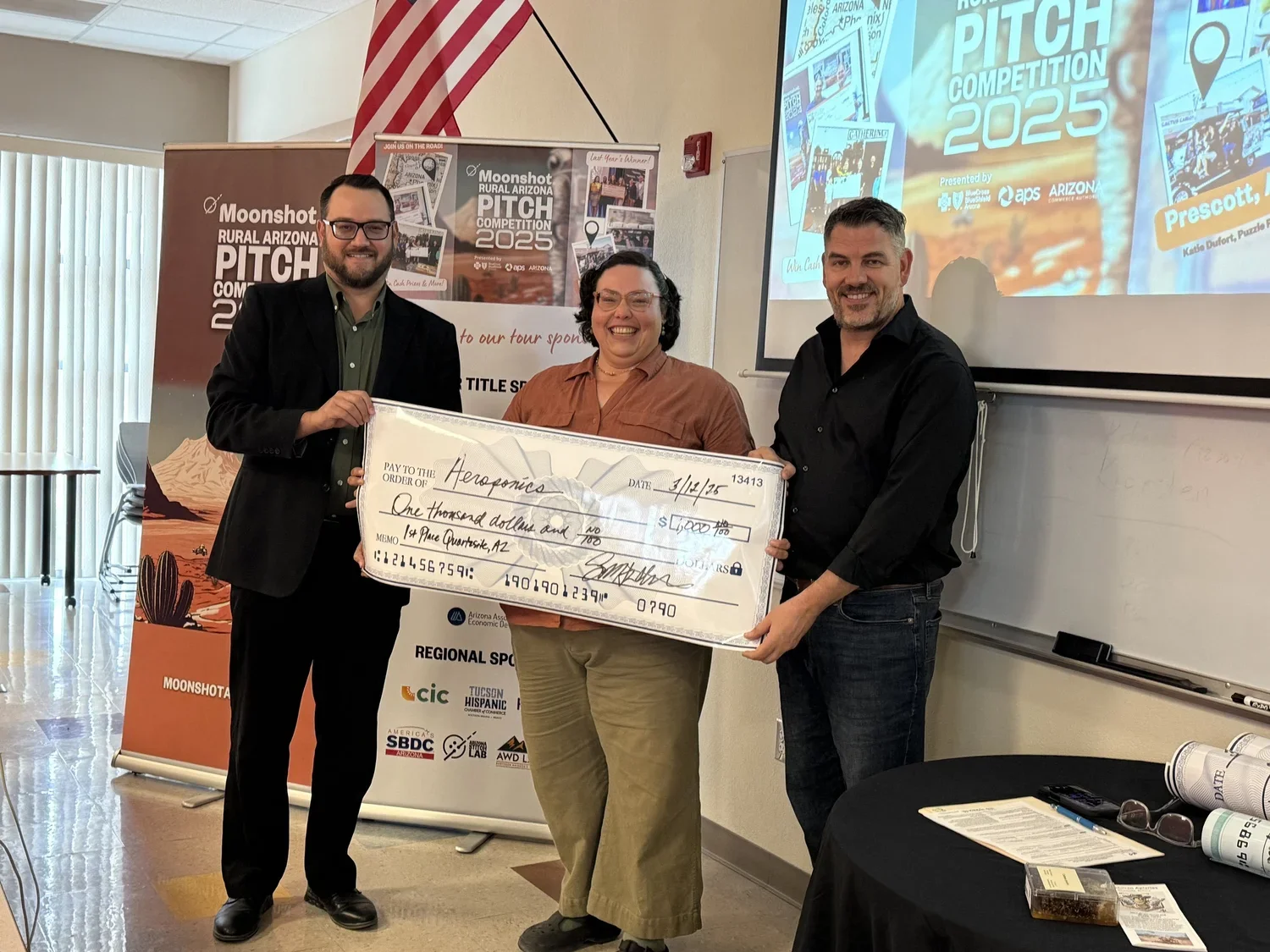 Three people are standing in front of a presentation slide and a banner, holding a large check. The check is made out to 'Aerospace' for one thousand dollars. The person on the left is a man in a black suit, the person in the middle is a woman with glasses, and the person on the right is a man in a black shirt. They are smiling and posing for the photo.