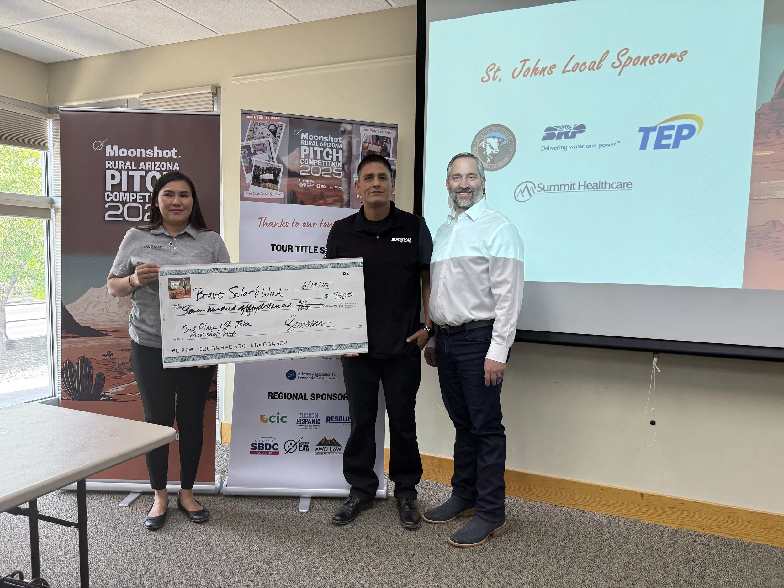 Three individuals stand together, with one holding an oversized check, at the Moonshot Rural Arizona Pitch Competition 2023 event, in front of a promotional banner and a presentation screen displaying sponsor logos.