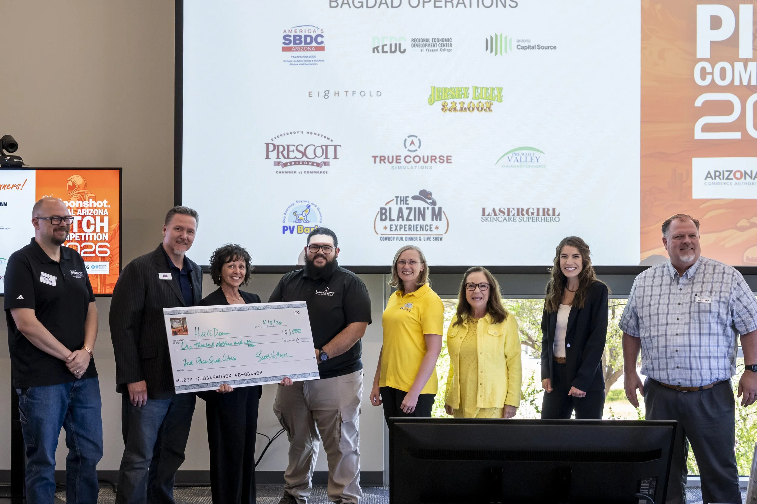 People standing in a row in an indoor setting, with a large screen behind them displaying logos of various organizations. The group includes five men and three women, some holding a large check. They are smiling and dressed in business casual attire.