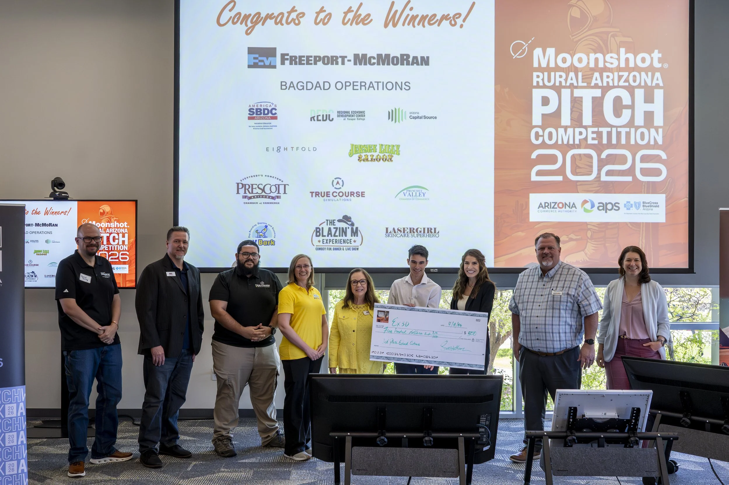 Group of eight people standing in a room, holding a large check, at the Moonshot Arizona Pitch Competition 2026. Behind them, a large presentation screen displays the winners and sponsor logos.