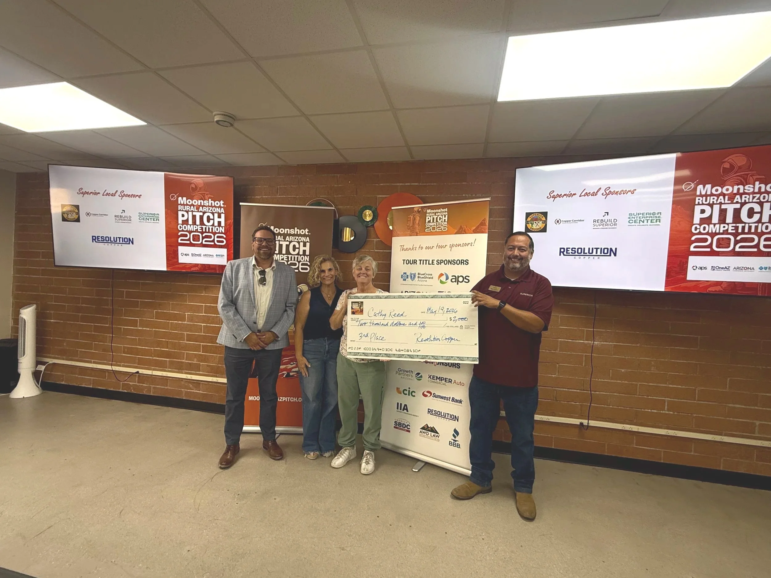 Three people standing together holding a large check during the Moonshot Rural Arizona Pitch Competition 2025. The person on the left is a tall man with dark hair and beard, wearing a black shirt. The woman in the middle has blond curly hair and is wearing a patterned dress. The woman on the right has dark hair tied back, wearing a beige blazer and blue jeans. Behind them are banners for the event and a brick wall.