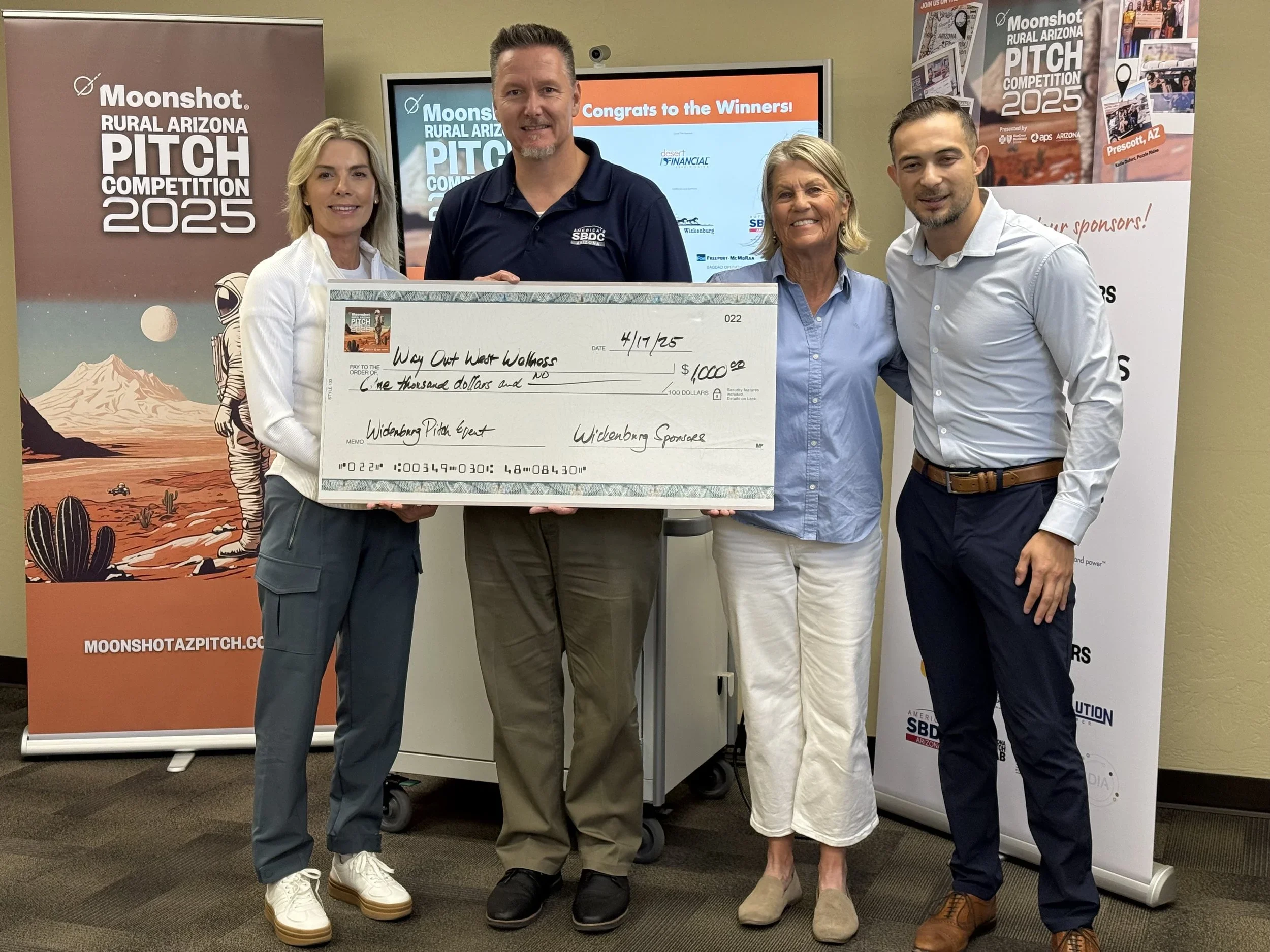 Four people standing indoors, holding a large check for one thousand dollars made out to Way Out West Walkers, at an event for Moonshot rural Arizona pitch competition. Behind them are banners for the event and a flat-screen monitor displaying congratulatory text.