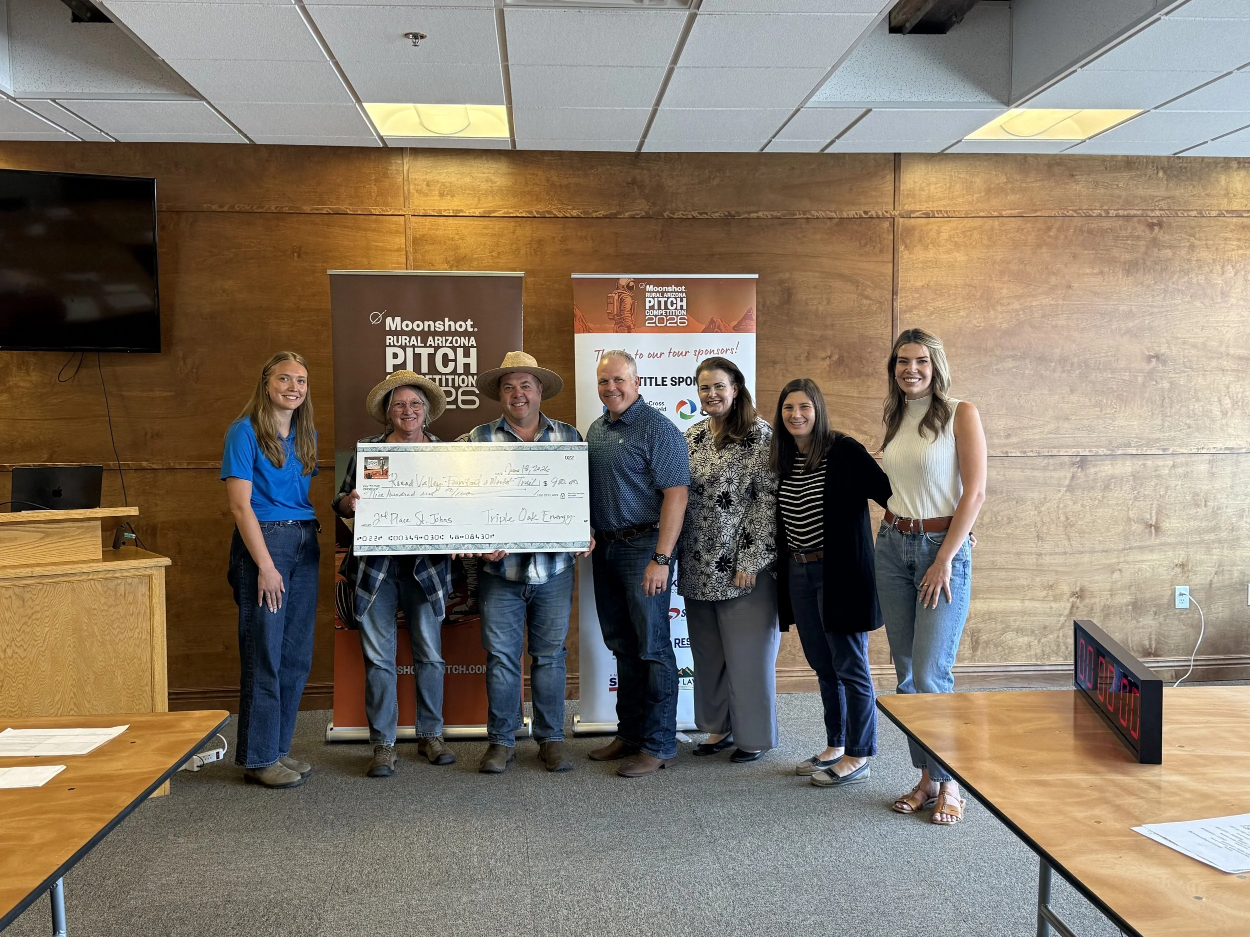 Three individuals stand together, with one holding an oversized check, at the Moonshot Rural Arizona Pitch Competition 2023 event, in front of a promotional banner and a presentation screen displaying sponsor logos.