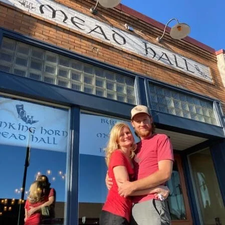 A couple in red shirts embracing in front of a building with a sign that reads 'Mead Hall.' The facade includes large windows and retro-style lights.
