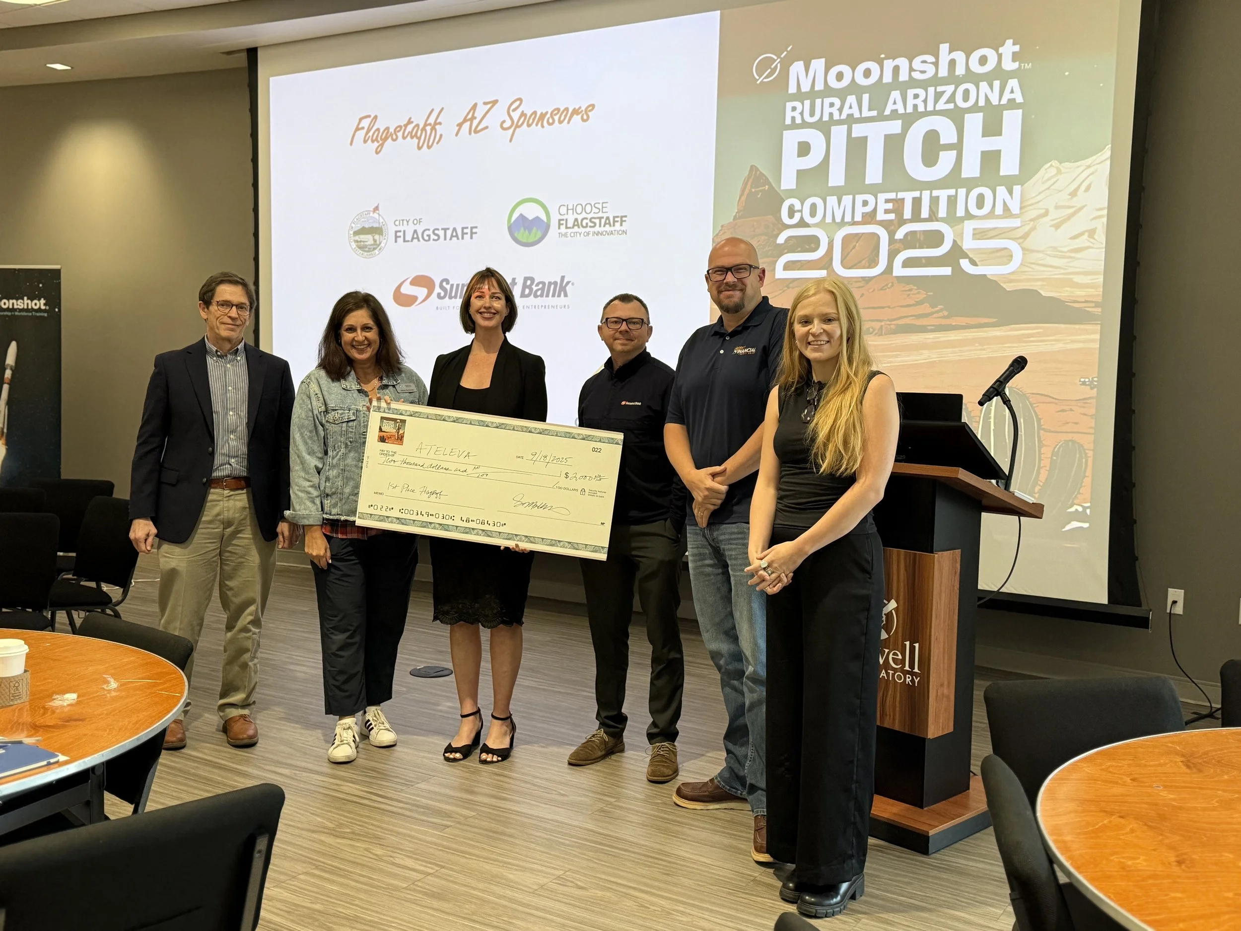 Group of six people standing on stage at the Moonshot Rural Arizona Pitch Competition 2025, holding a giant check, with a presentation screen in the background displaying event sponsors and logos.