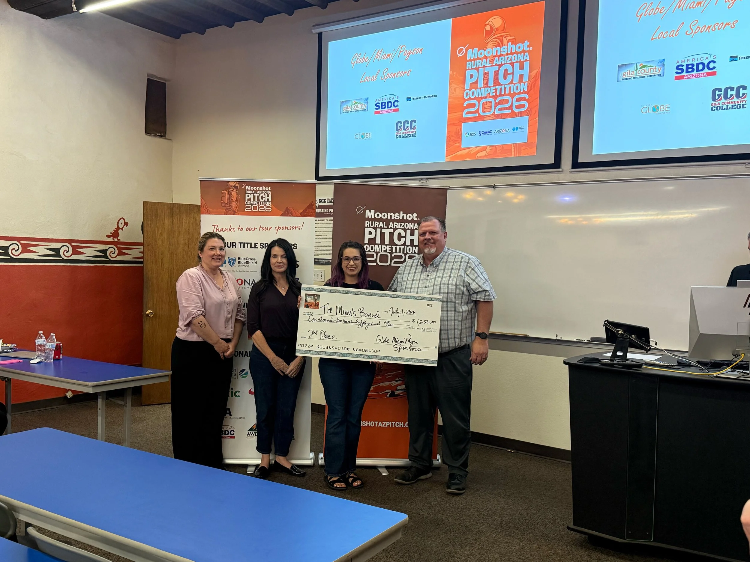 Group of six people standing indoors holding a large check for $1,000, celebrating an award at the Moonshot Rural Arizona Pitch Competition 2025, with banners and sponsor signs in the background.