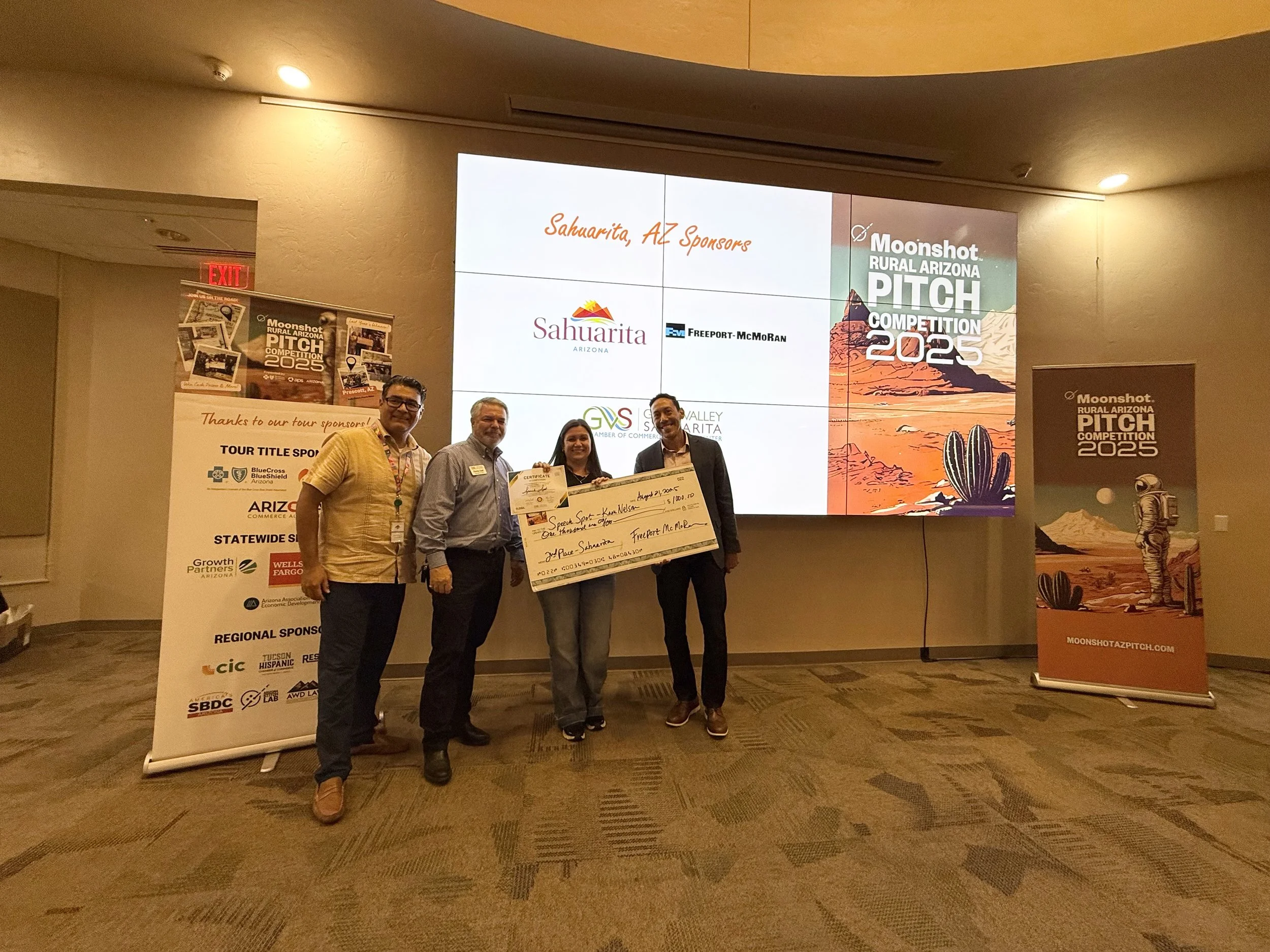 A group of four people standing on stage at an awards ceremony, holding a large check with the words 'Speech Kwan Nelson' and '2nd Place Sahuarita' written on it. Behind them, there are two banners and a large screen displaying the logo and details of the Moonshot Rural Arizona Pitch Competition 2025, featuring desert landscape art and an astronaut.