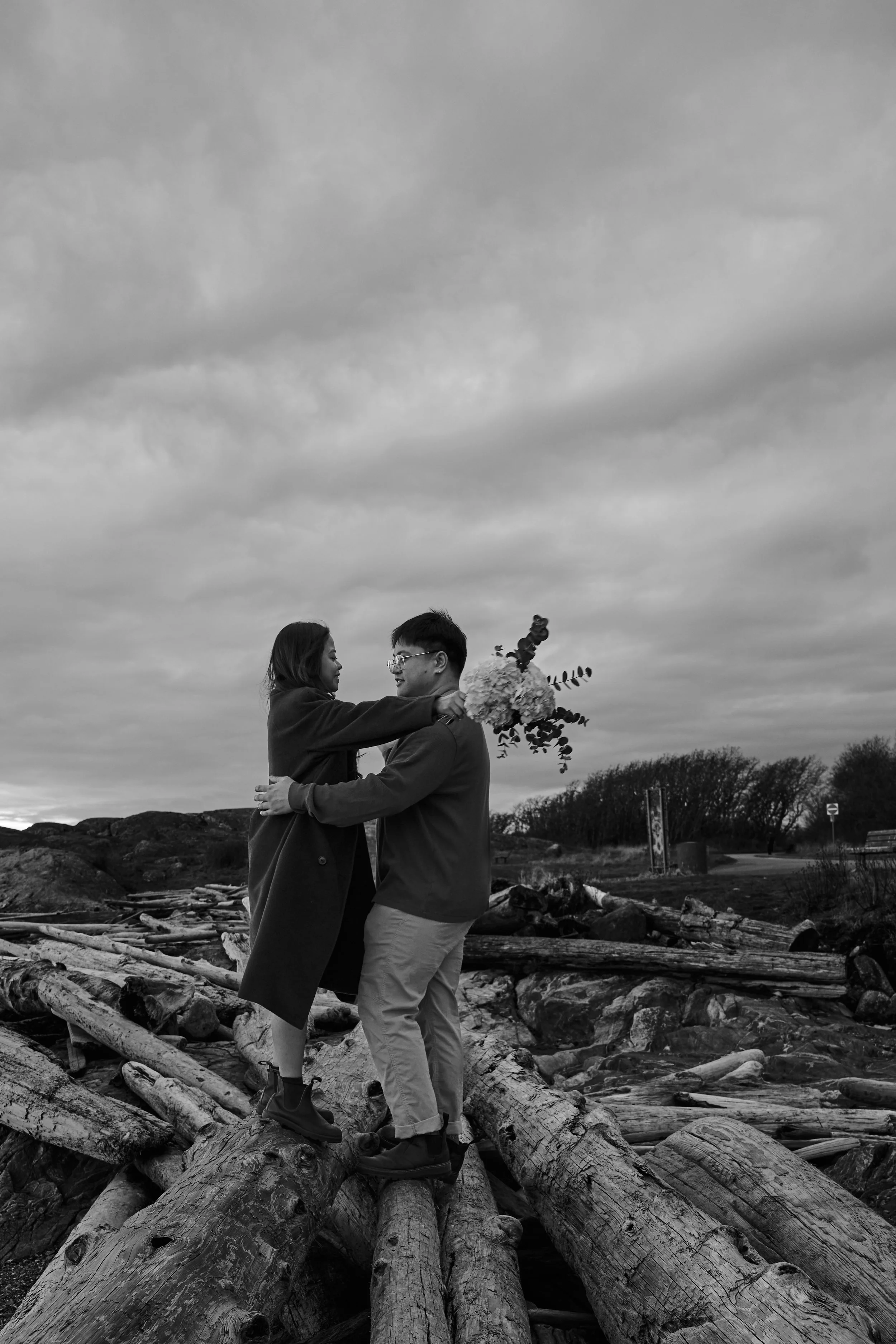 A couple embracing on a pile of driftwood with overcast skies, in a coastal or natural setting. One person holds a bouquet of flowers.