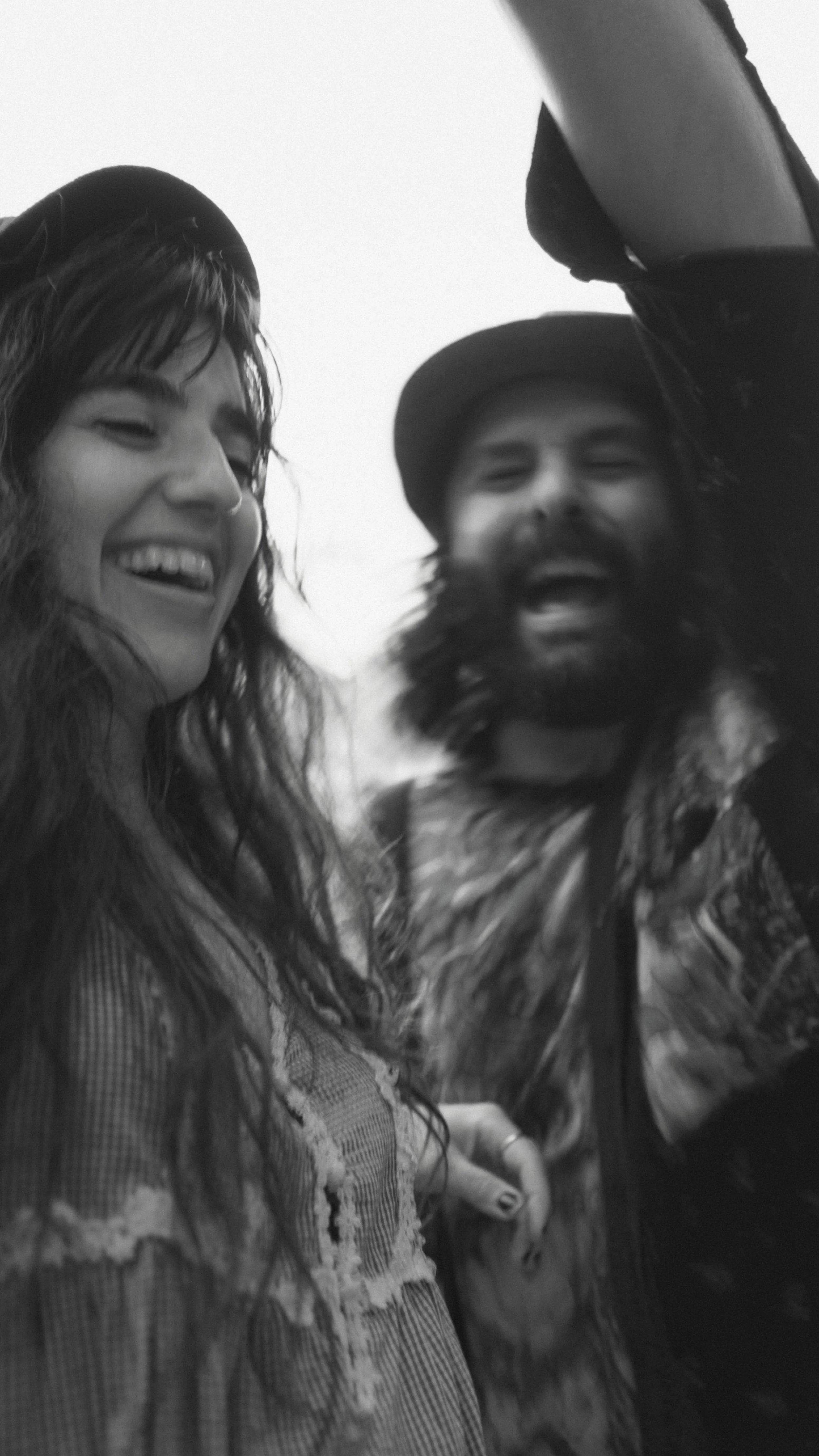 Black and white photo of two people smiling and laughing, captured candidly. One has long hair, and the other has a beard and is wearing a hat. The mood is joyful and dynamic.