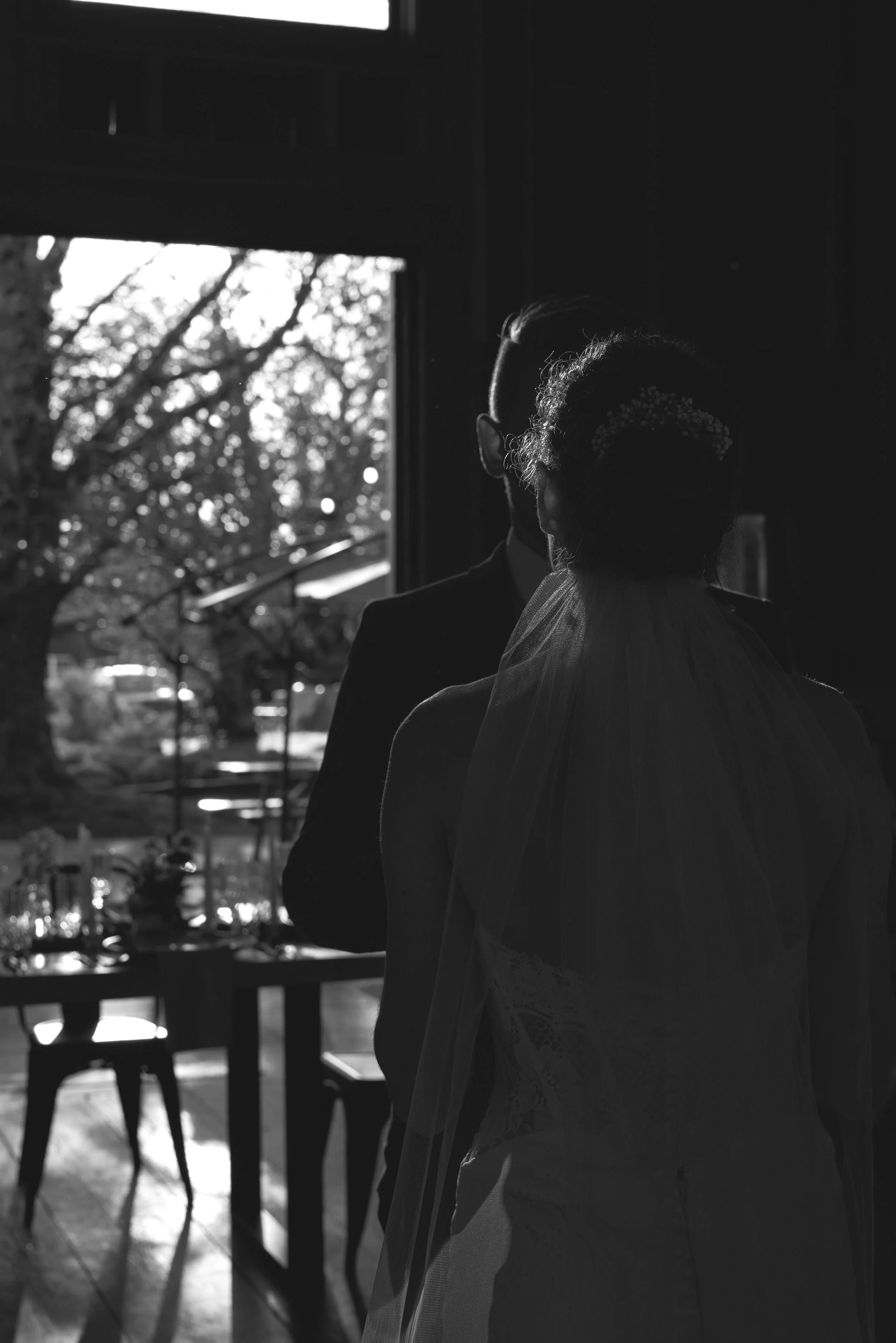 Silhouetted couple standing indoors, woman in bridal attire, sunlight streaming through window, tree outside.