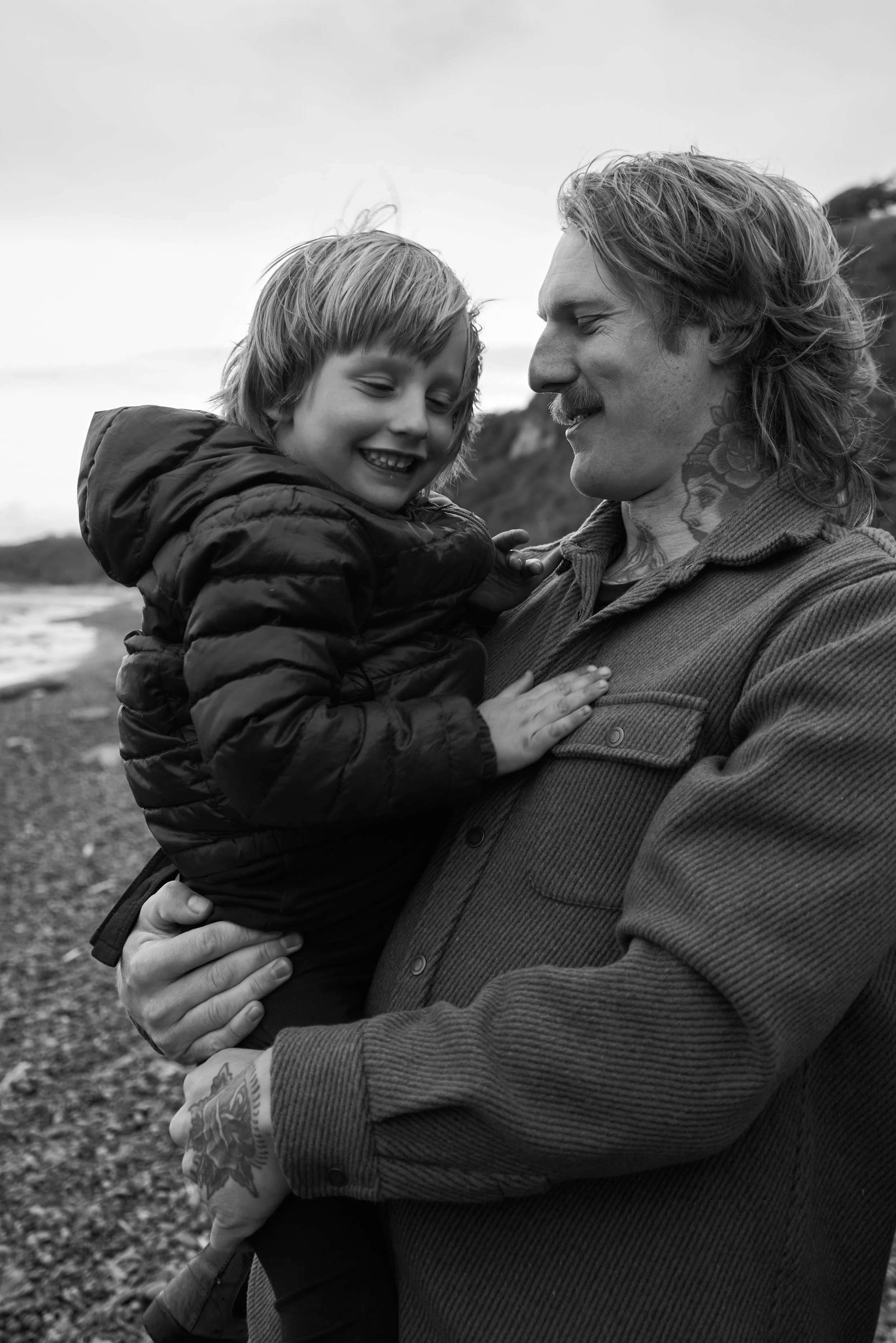 Black and white photo of a man holding a smiling child by the beach. The man has tattoos and long hair. Both are looking at each other and smiling.