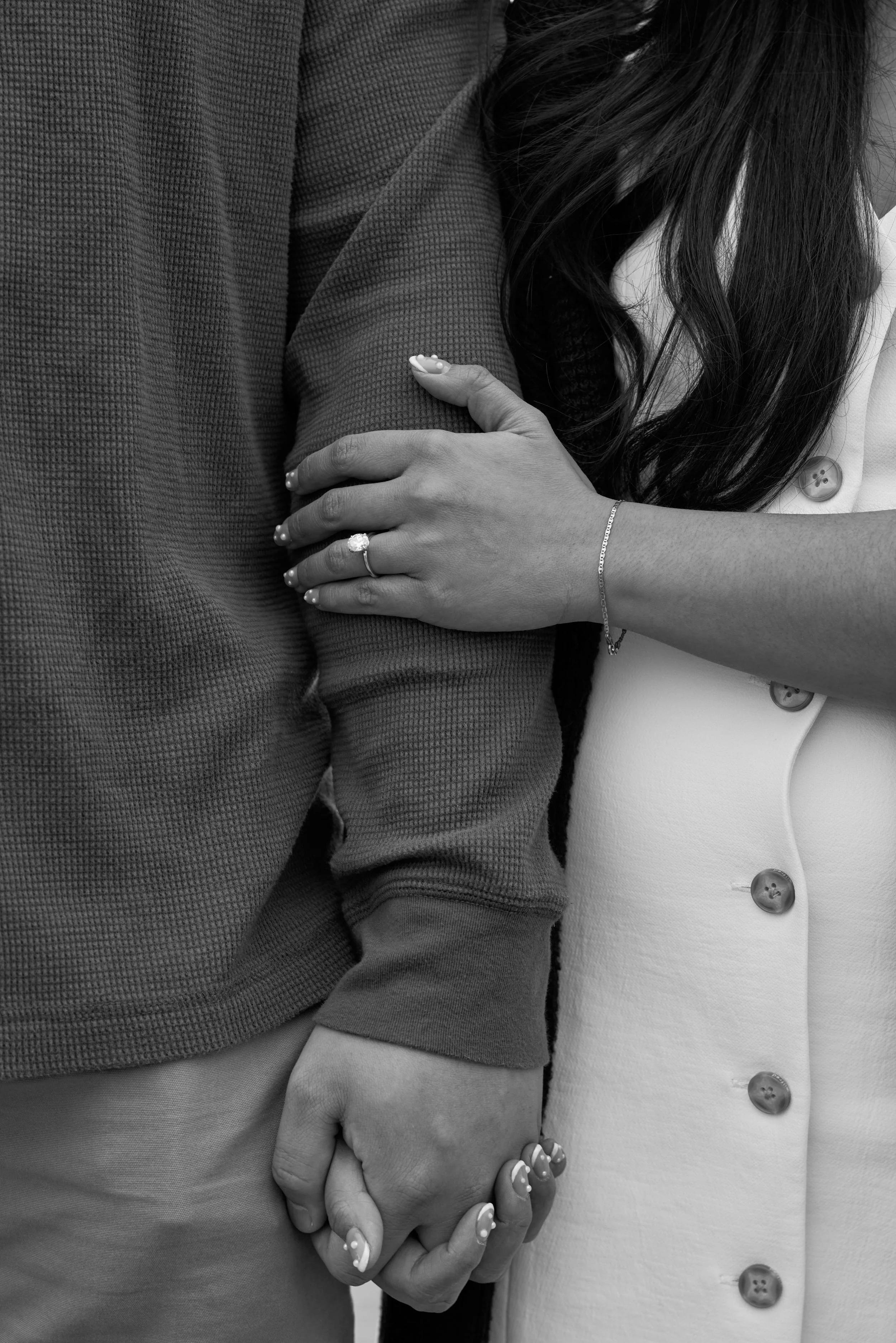 Black and white image of a couple holding hands. The woman wears a ring and has her arm linked with the man's arm.