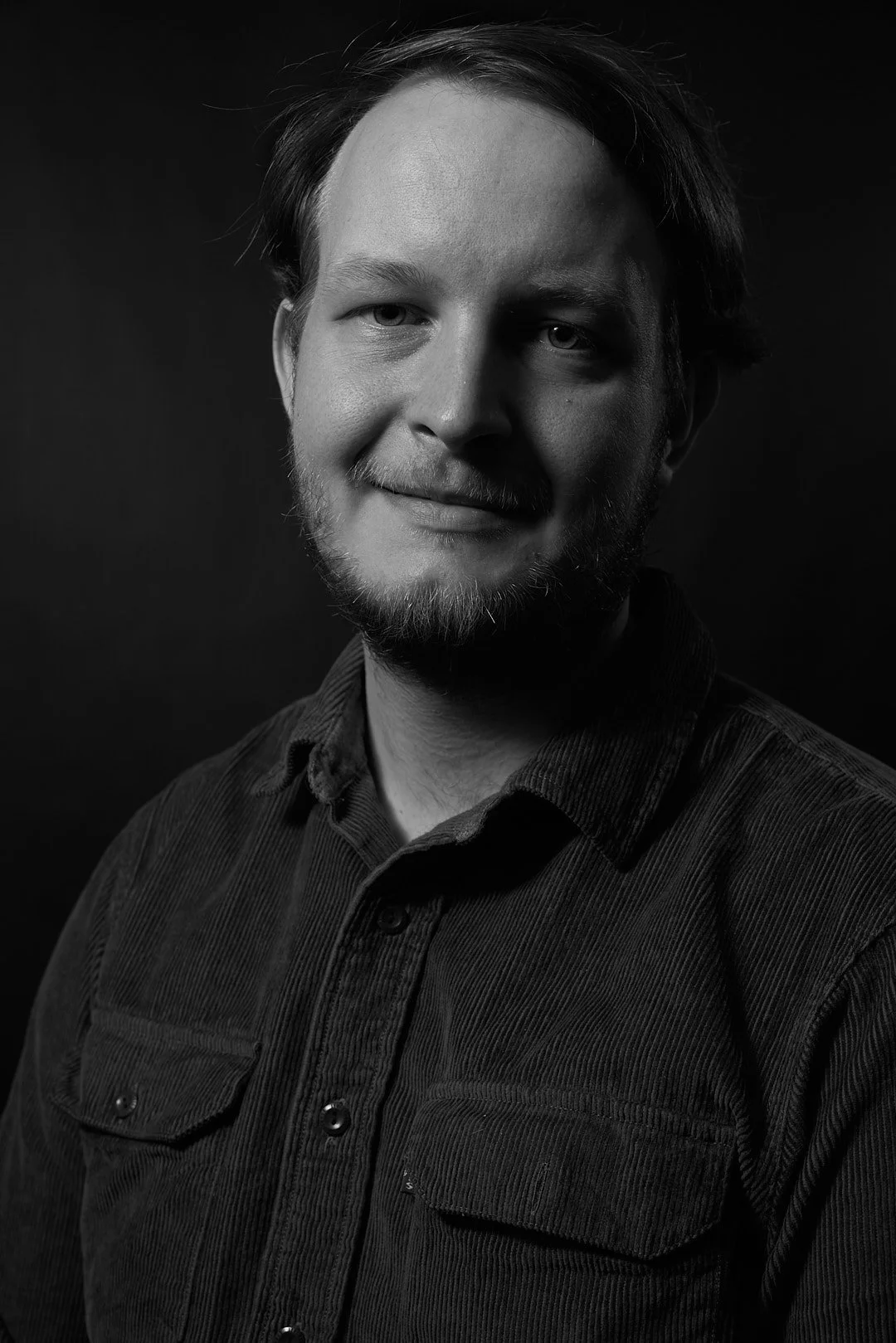 Black and white portrait of a man with a beard, wearing a dark button-up shirt, looking towards the camera.